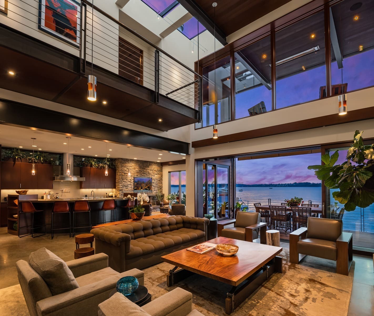 A spacious, modern living room features a mix of wood and stone elements. It includes a plush sofa, armchairs, coffee table, and a dining area. Large glass doors open to a scenic balcony with water views at sunset. Contemporary decor and pendant lights add elegance.