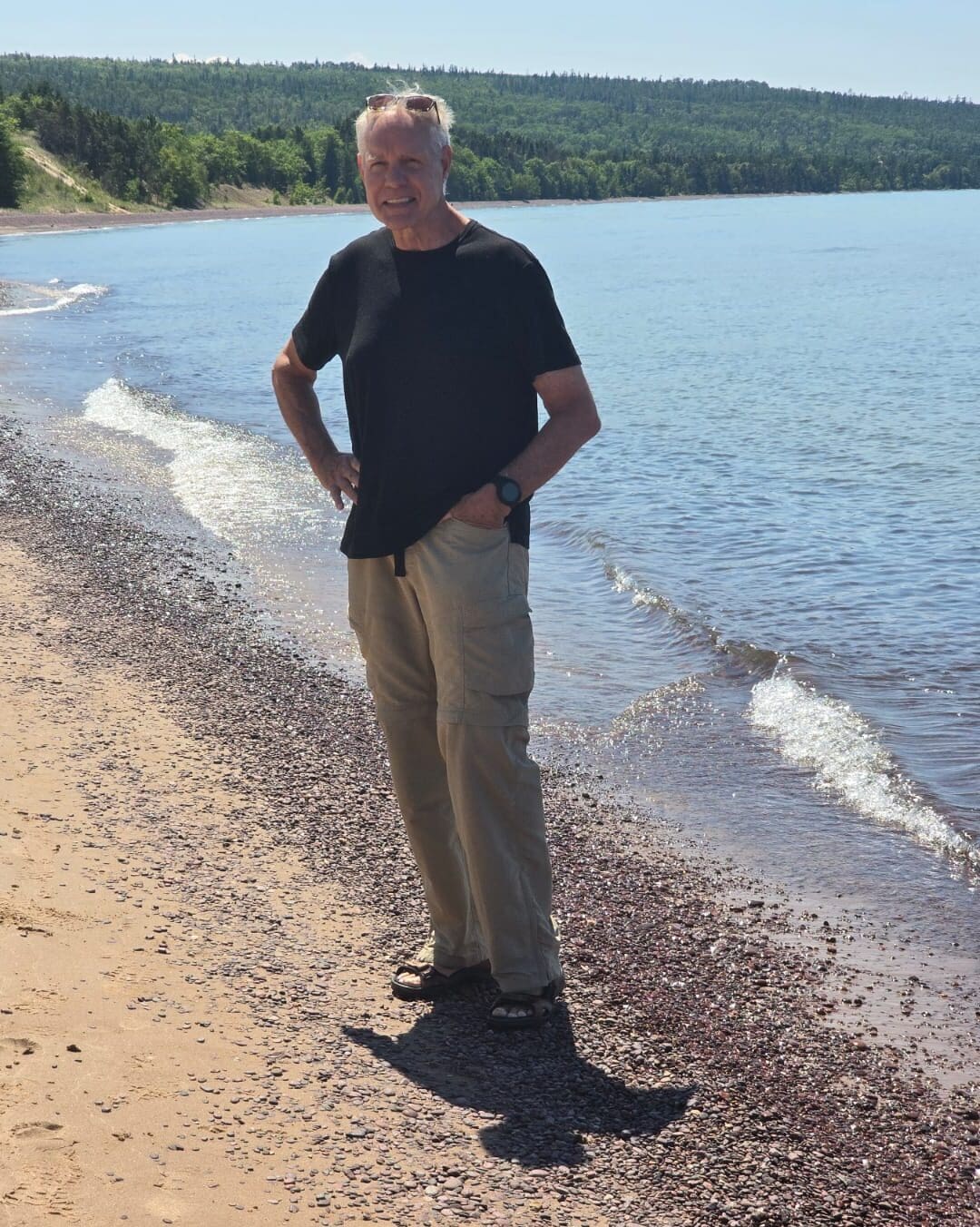 Man standing on a sandy beach by a tranquil lake, surrounded by lush greenery in the background, showcasing a relaxed summer vibe, ideal for outdoor living in the Pacific Northwest.
