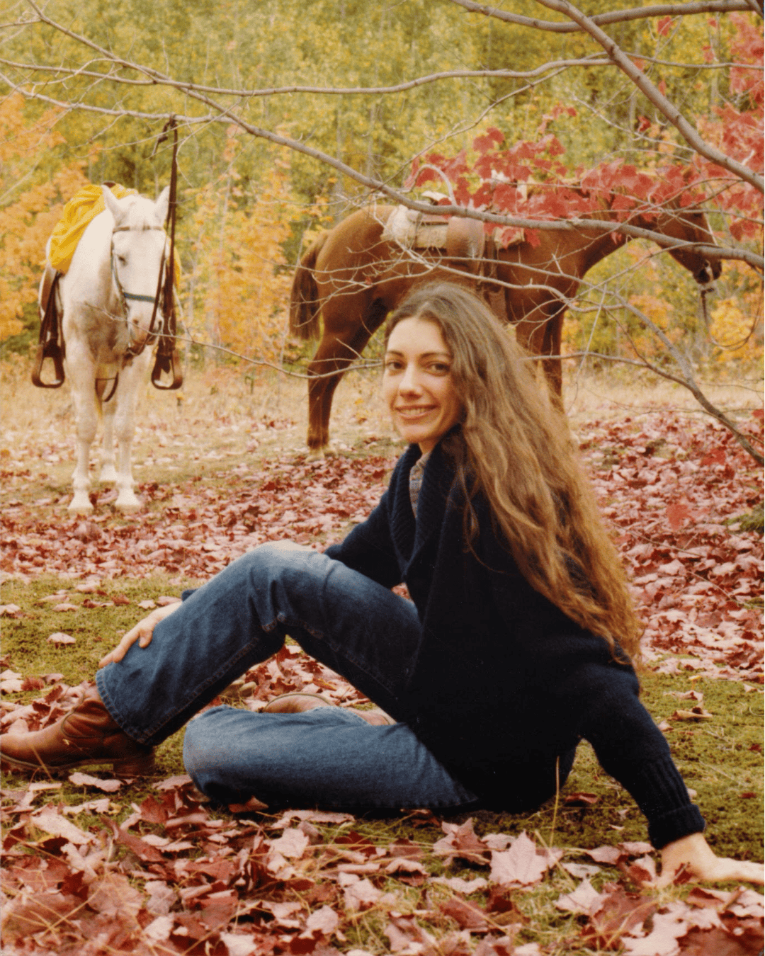 A woman sitting on a bed of colorful autumn leaves with two horses in the background, set in a serene outdoor landscape typical of the Pacific Northwest. The scene captures a peaceful, rustic vibe, highlighting the natural beauty of fall.