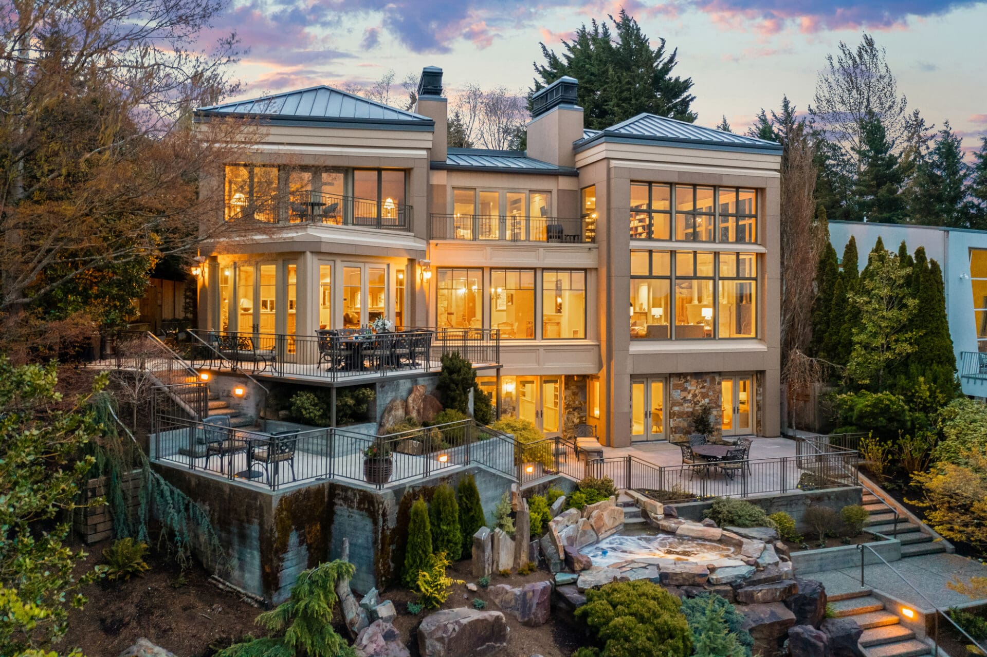 Stunning modern exterior of a luxury home in the Pacific Northwest, featuring expansive glass windows, elegant outdoor seating, and beautifully landscaped gardens, illuminated at twilight.