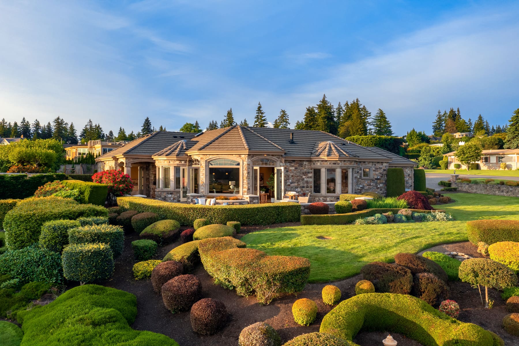 Elegant exterior view of a luxury home surrounded by meticulously landscaped gardens in the Pacific Northwest, showcasing a blend of stone architecture and lush greenery under a clear blue sky.