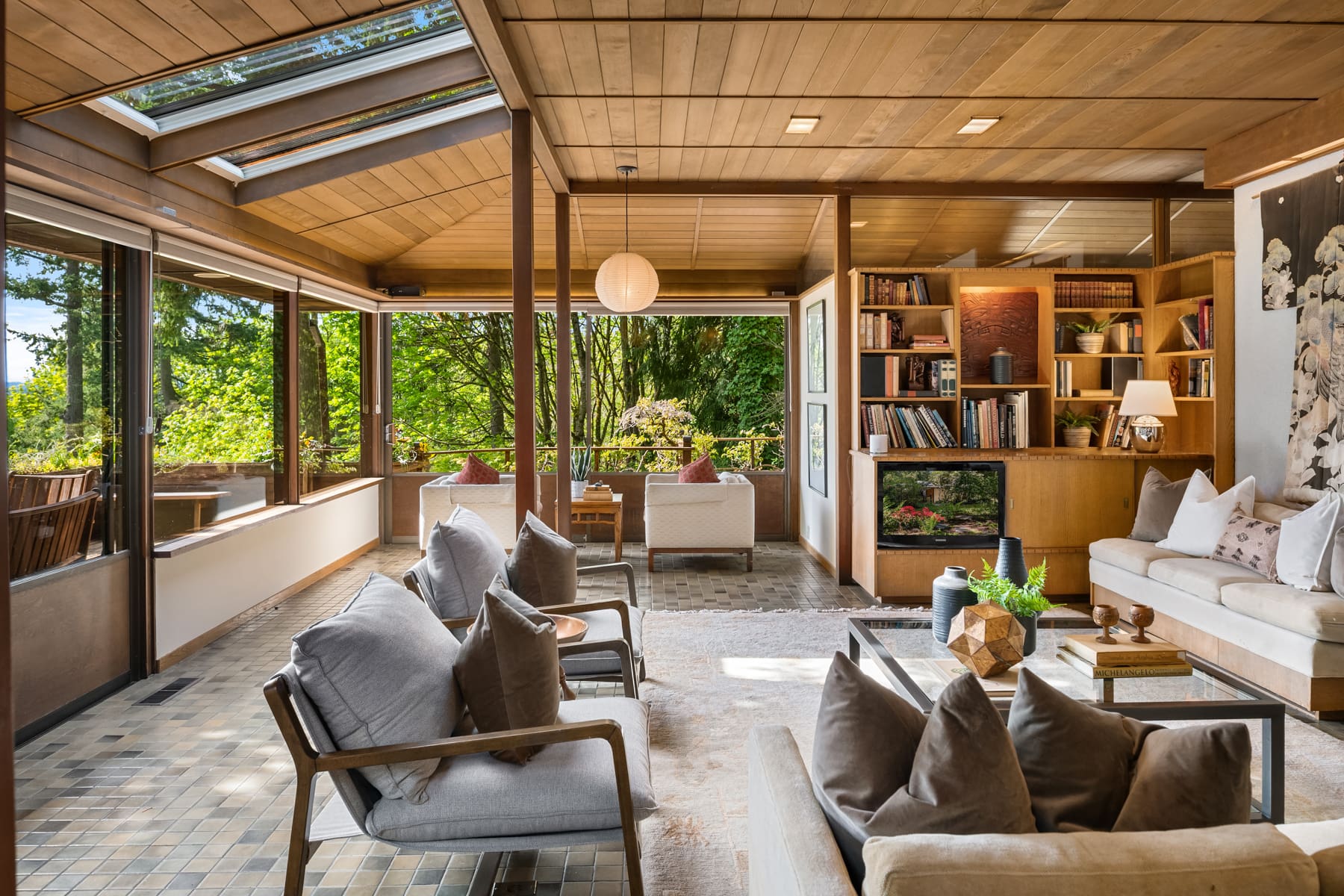 Modern living room with natural wood accents and large windows showcasing lush greenery, featuring cozy seating and a stylish bookshelf, located in the Pacific Northwest.