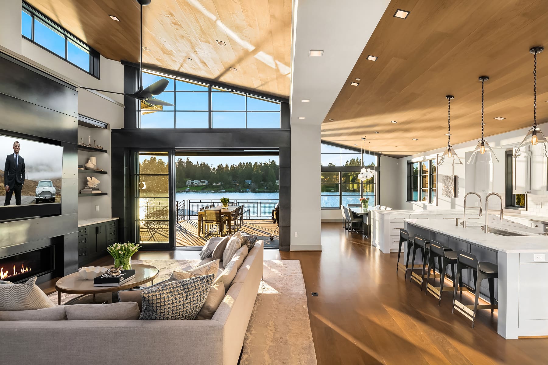 Modern waterfront living room and kitchen in a Pacific Northwest home, featuring large windows, a sleek design with wood accents, and a cozy ambiance, showcasing views of the water and outdoor dining area.