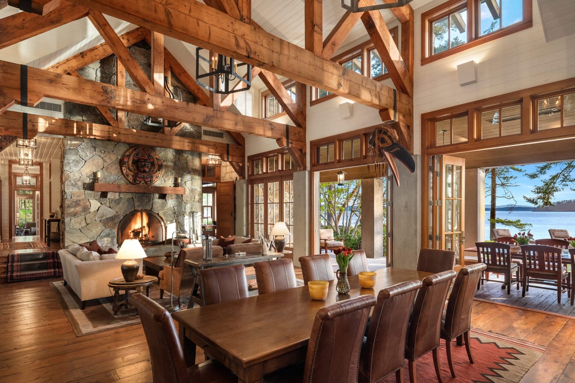 A spacious, rustic living and dining area with exposed wooden beams, a large stone fireplace, and high ceilings. Perfect for real estate photoshoots, the room features a large wooden dining table with chairs, cozy seating areas, and expansive windows offering HDR views of the lake and surrounding trees.