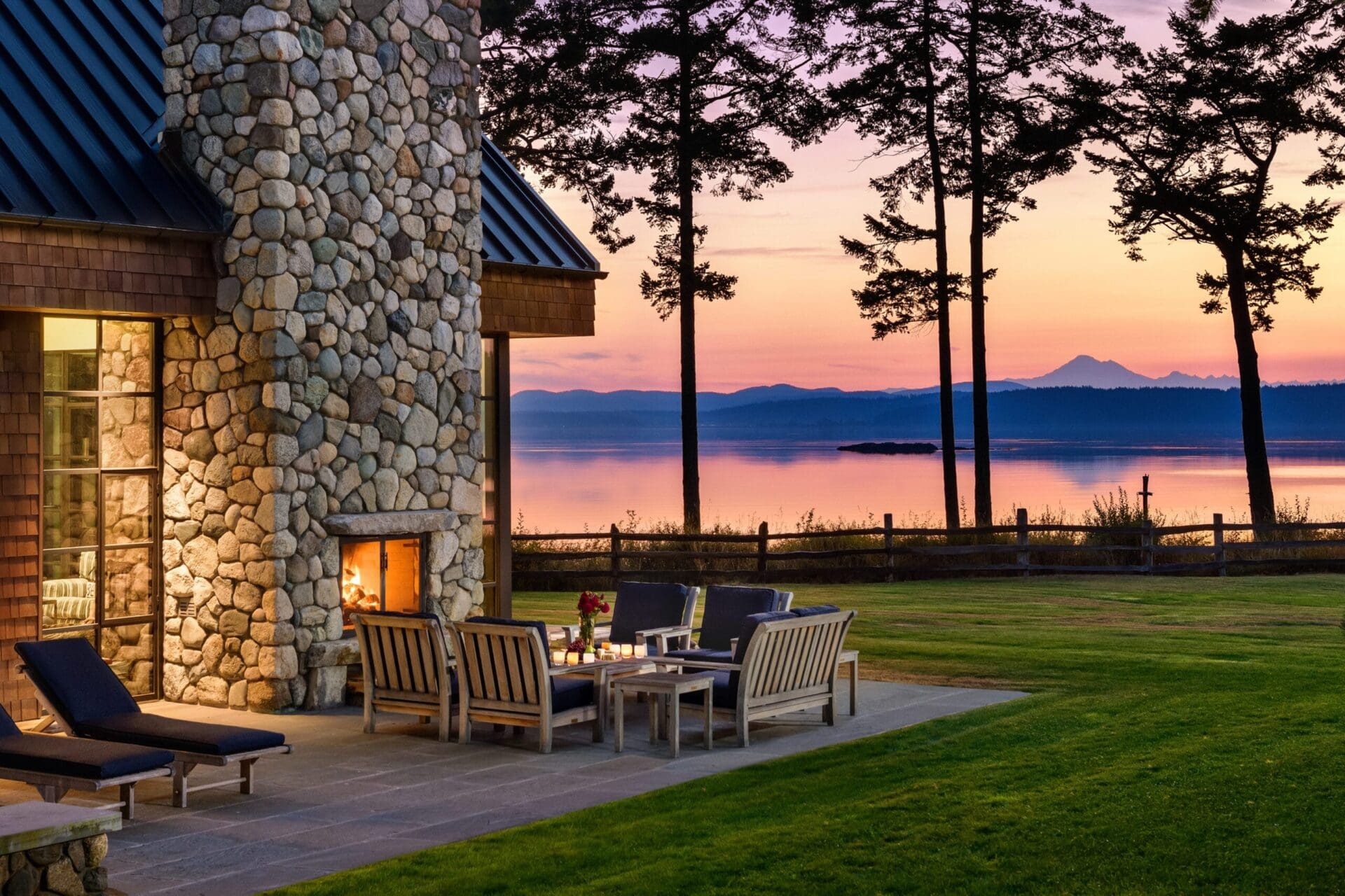 A serene lakeside patio at sunset, featuring a stone fireplace, cushioned wooden chairs around a coffee table with a flower vase, and lounge chairs. Tall trees frame the view of the calm lake and distant mountains against a colorful sky.
