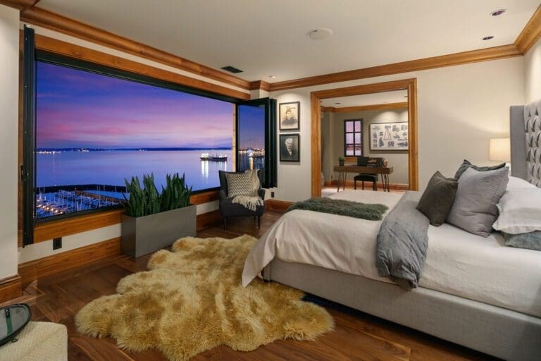 A cozy bedroom with a large open window facing a peaceful harbor at sunset. The room features a plush rug, modern furniture with grey and white bedding, potted plants, and wooden accents, creating a warm and inviting atmosphere. Photos and pictures adorn the walls. Seattle Luxury Real Estate Photography