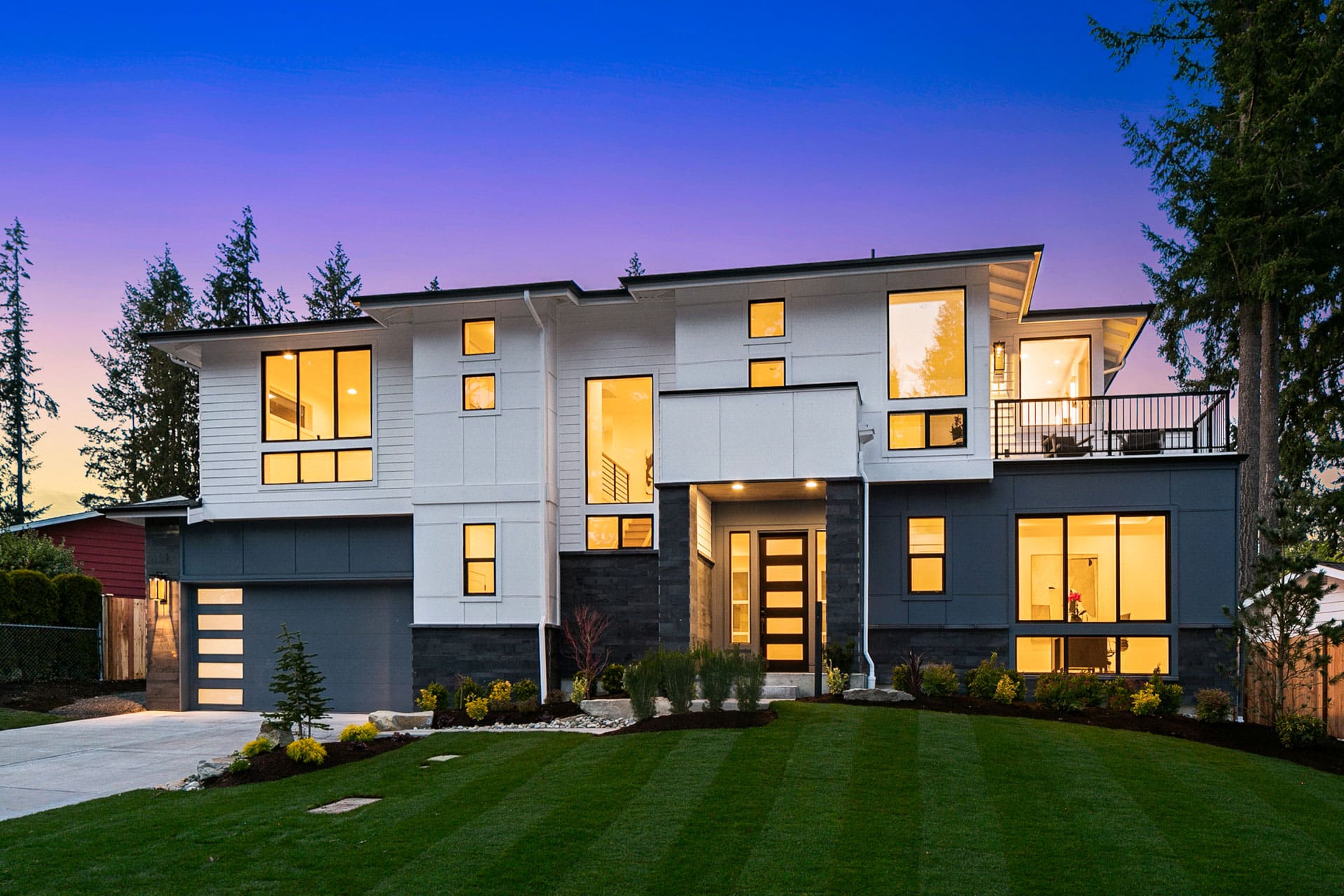 westside real estate photography A modern two-story house with a well-manicured lawn and a paved driveway. The house features large windows, a contemporary design with a mix of white and dark exterior materials, and a prominent front entrance. Tall trees and a twilight sky are in the background.