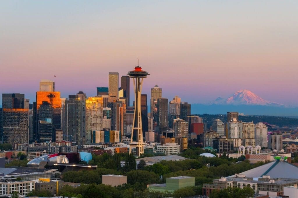 Aerial view of downtown Seattle at sunset with the Space Needle prominently in the center. Skyscrapers glow with warm light against a pastel sky, and Mount Rainier is visible in the background. Greenery and urban buildings are seen at the base.