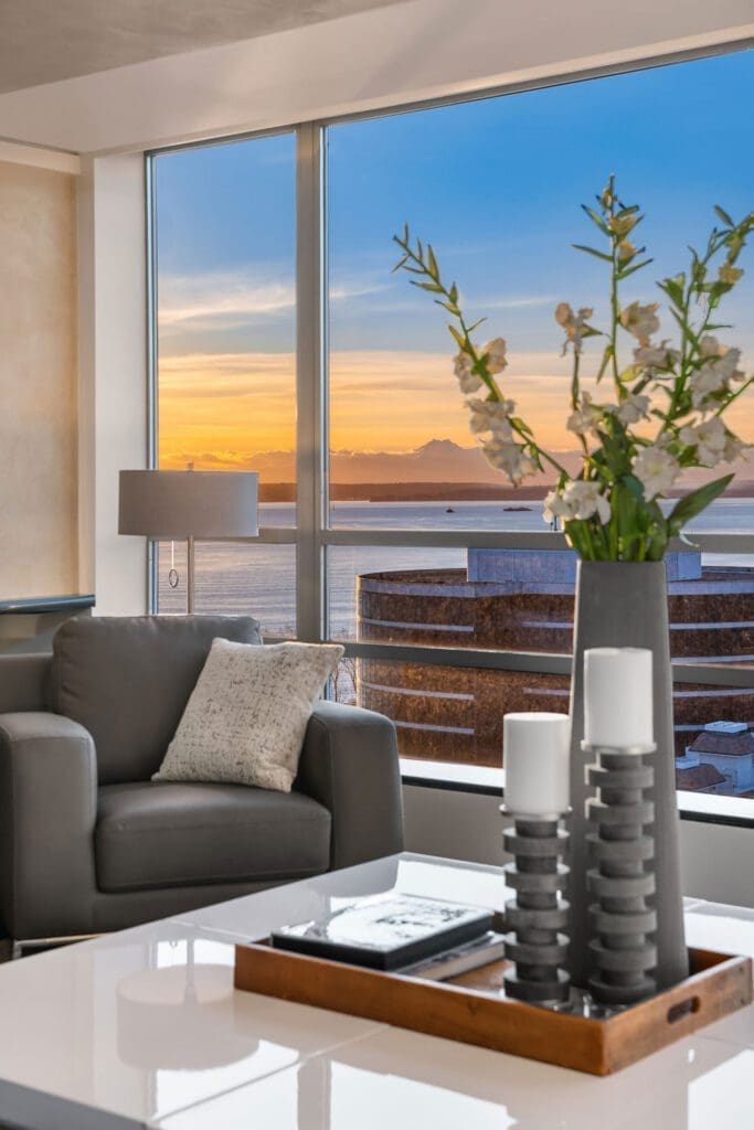 A modern living room with a gray armchair, a pillow, and a side lamp in front of large windows showing a picturesque sunset over the ocean. A white coffee table holds a tray with candles, a magazine, and a tall vase with white flowers.