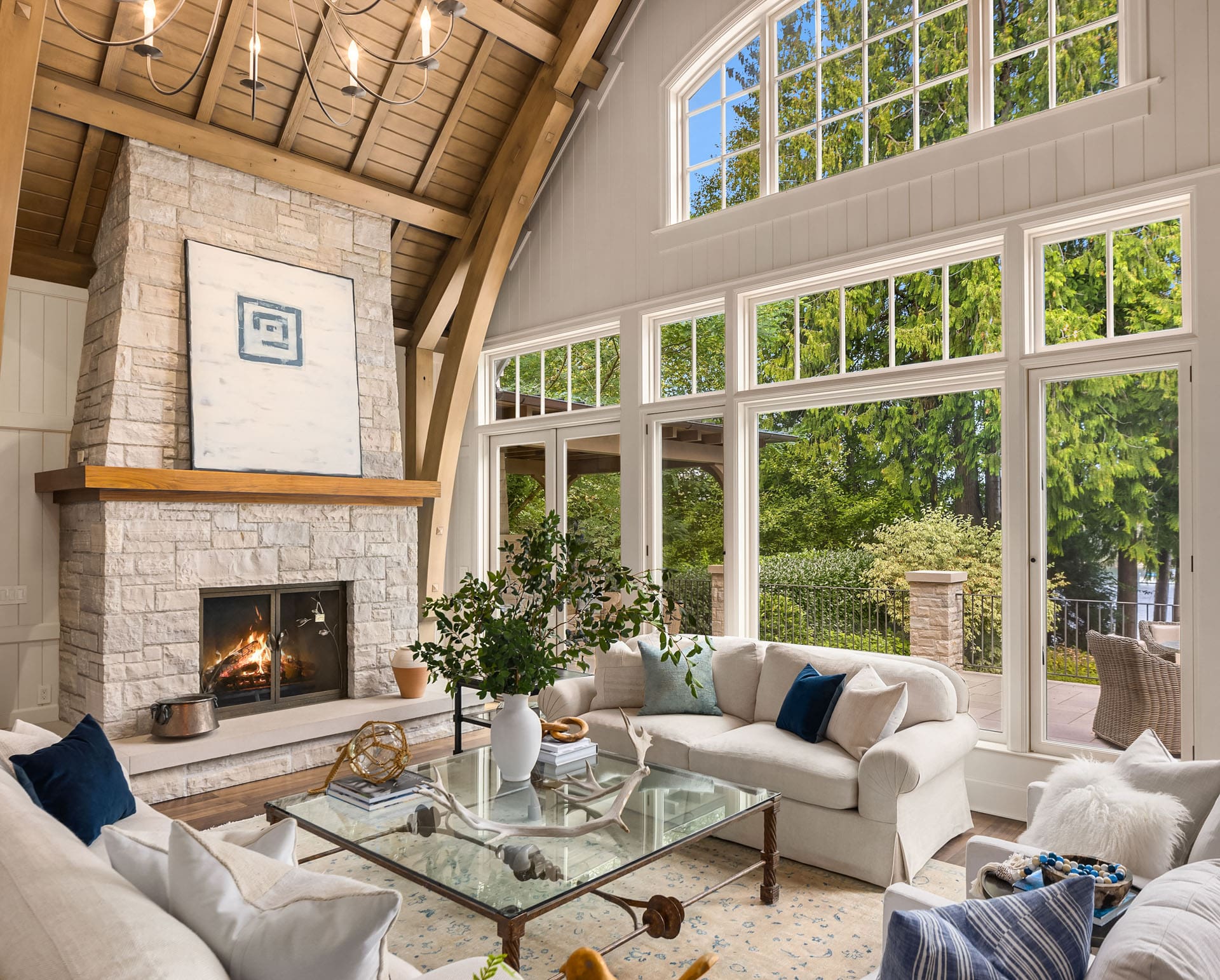 Island Real Estate Photography A cozy, elegantly designed living room features large windows, wooden beams, a stone fireplace, and white furnishings. A glass coffee table and greenery in a vase add a touch of nature. Outside, a lush green landscape is visible under a bright blue sky.