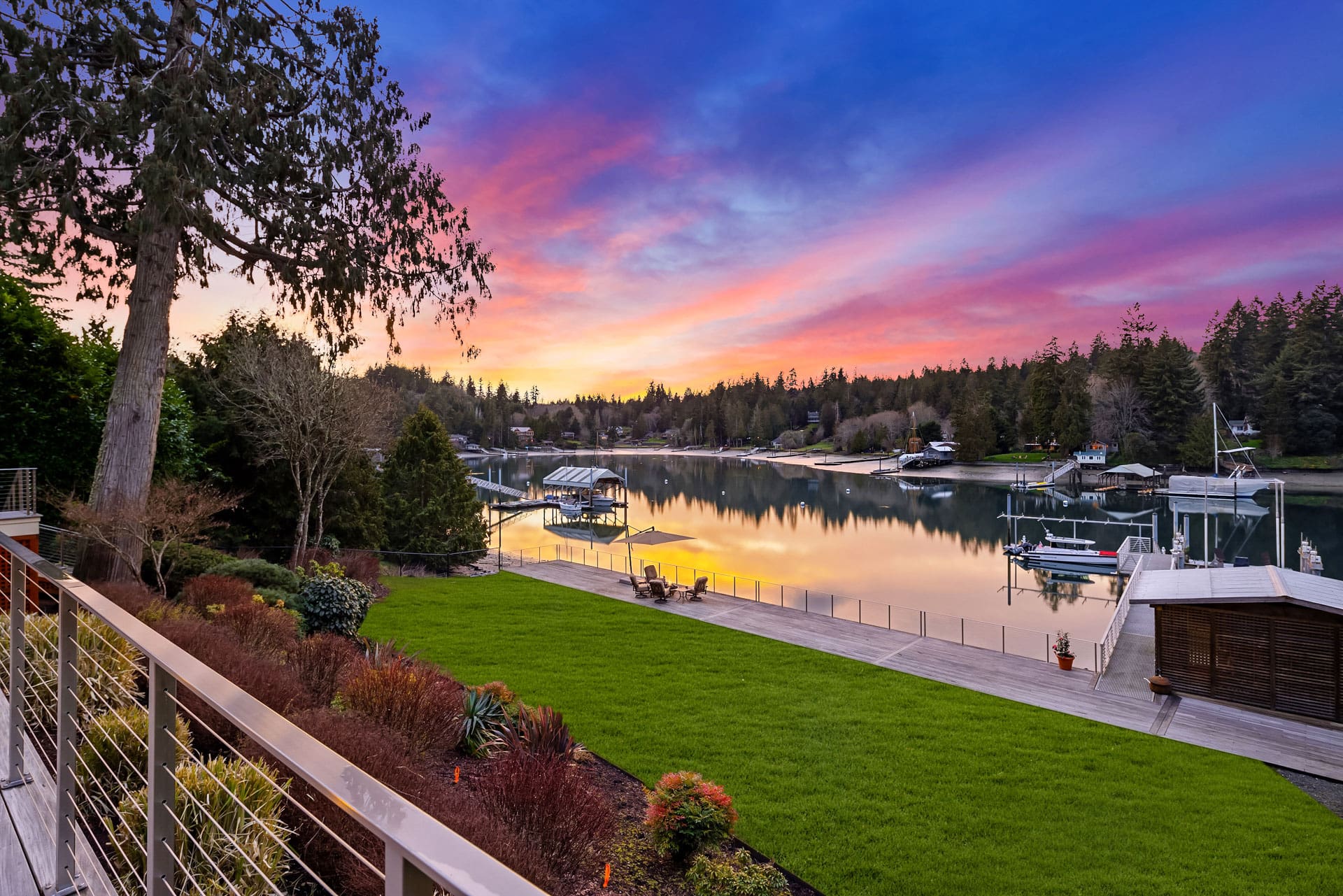 A tranquil lakeside scene at sunset featuring a well-maintained garden and a modern dock leading to the water. The sky is vibrant with pink, purple, and orange hues, reflecting on the calm water. Surrounded by tall trees, the area feels serene and picturesque.