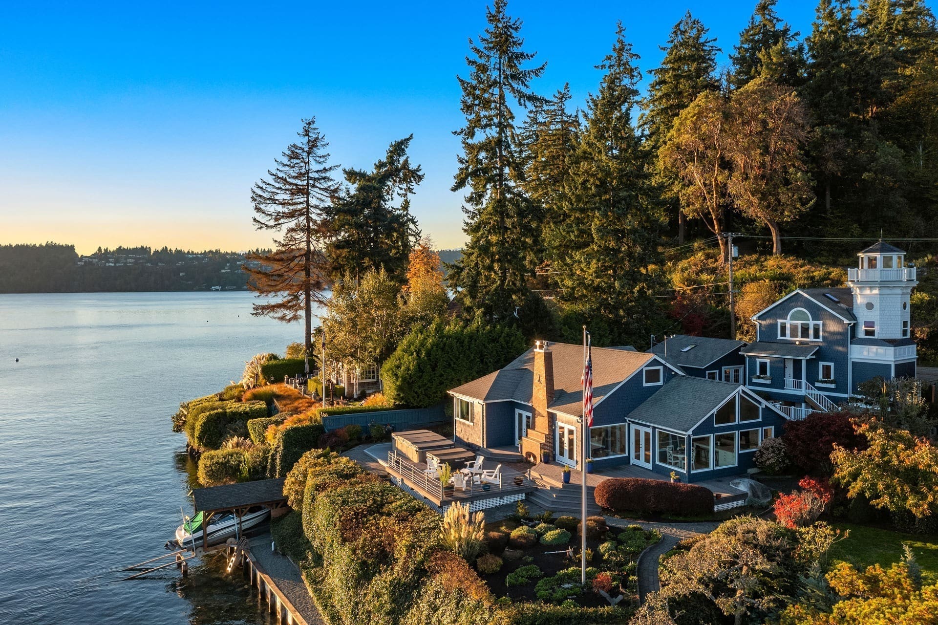 Southend Real Estate Photography Aerial view of a lakeside property with a large house surrounded by lush greenery and colorful landscaping. The house has multiple stories, several large windows, and a flagpole visible in the yard. A small dock extends into the calm lake, reflecting the clear blue sky.