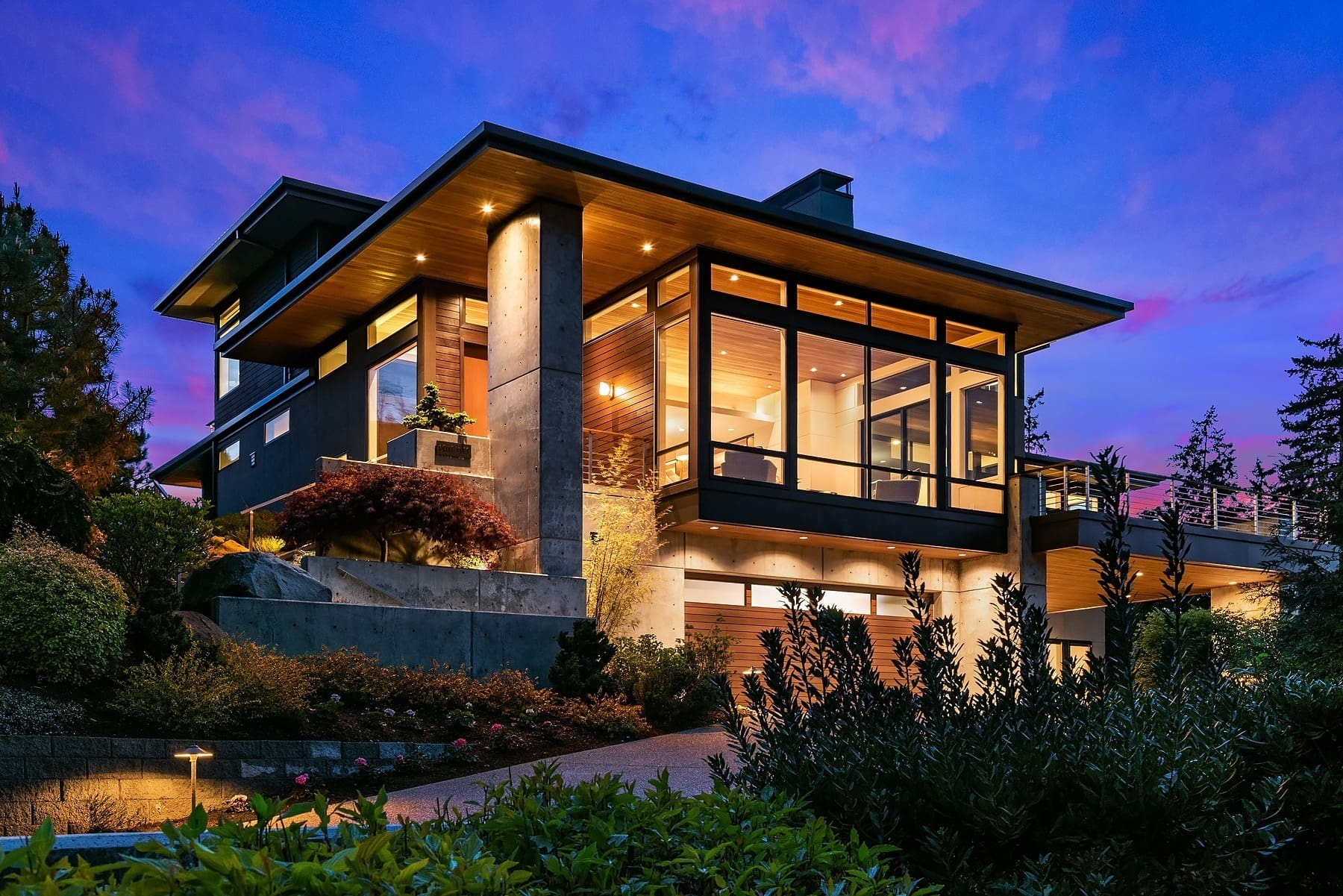 Greater Seattle Real Estate Photography Modern two-story house illuminated at dusk with large glass windows and wooden accents. The home features contemporary architecture with clean lines and concrete elements. Surrounded by lush greenery and strategically placed outdoor lighting.