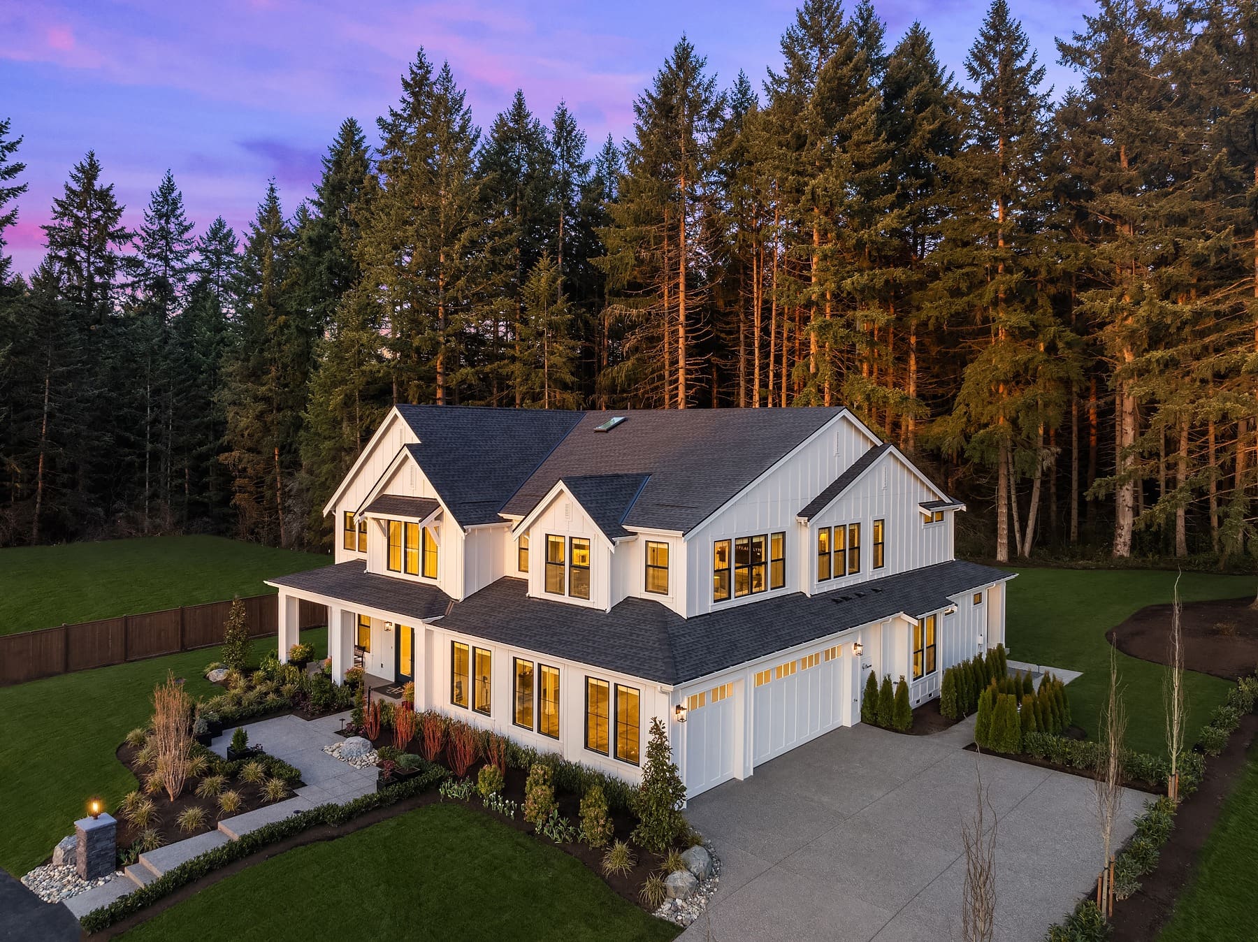 A modern two-story white house with large windows sits in a lush, landscaped yard surrounded by tall evergreen trees. The spacious driveway leads to a three-car garage. The sky is tinted purple and pink, indicating dusk or dawn. An open green lawn is visible on the left.