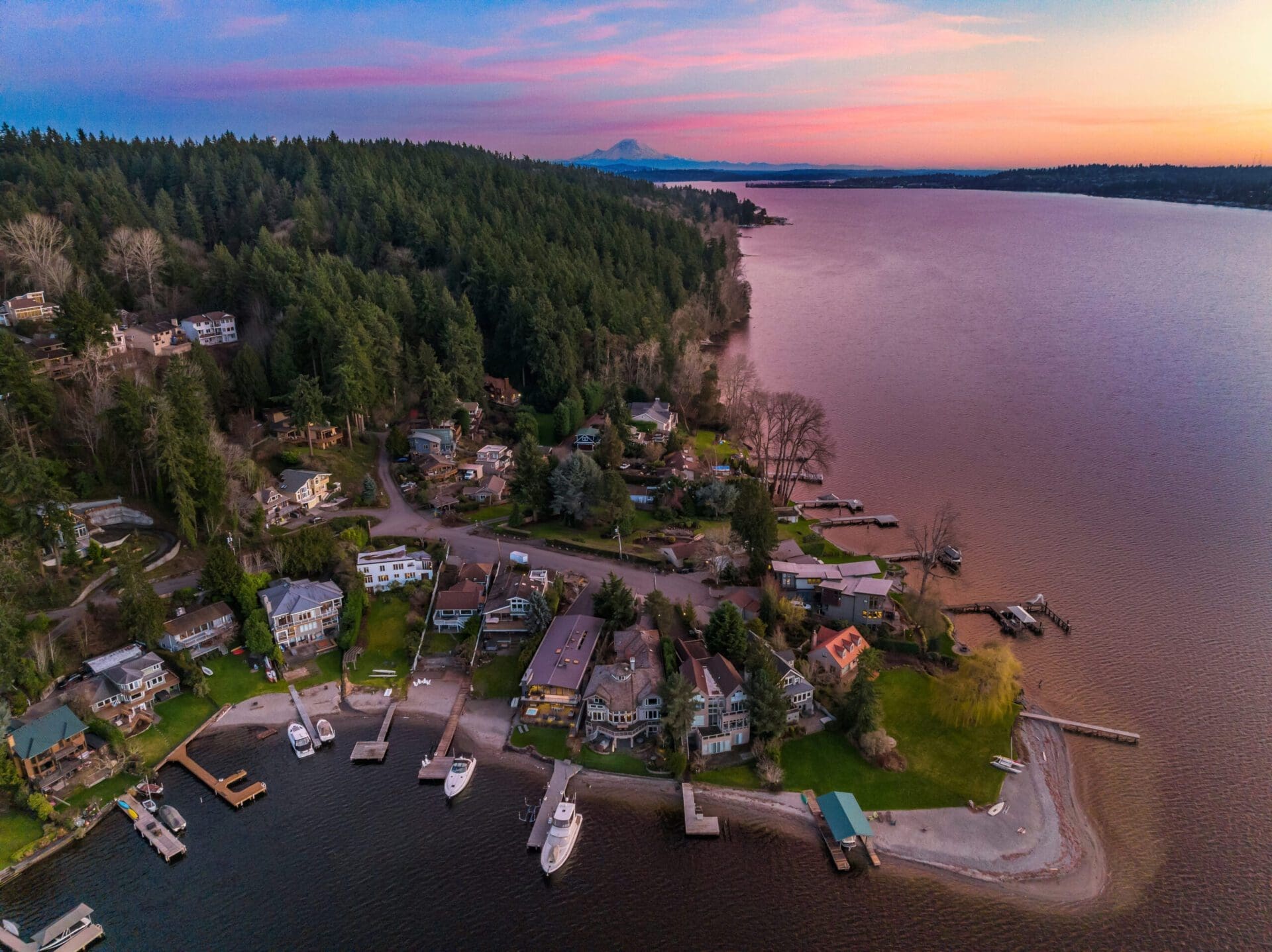 An aerial view of a lakeside residential area at sunset, with houses and docks lining the shore. Tall trees surround the homes, and a forest extends into the distance. The sky is painted with hues of pink, purple, and blue, and a mountain is visible on the horizon.