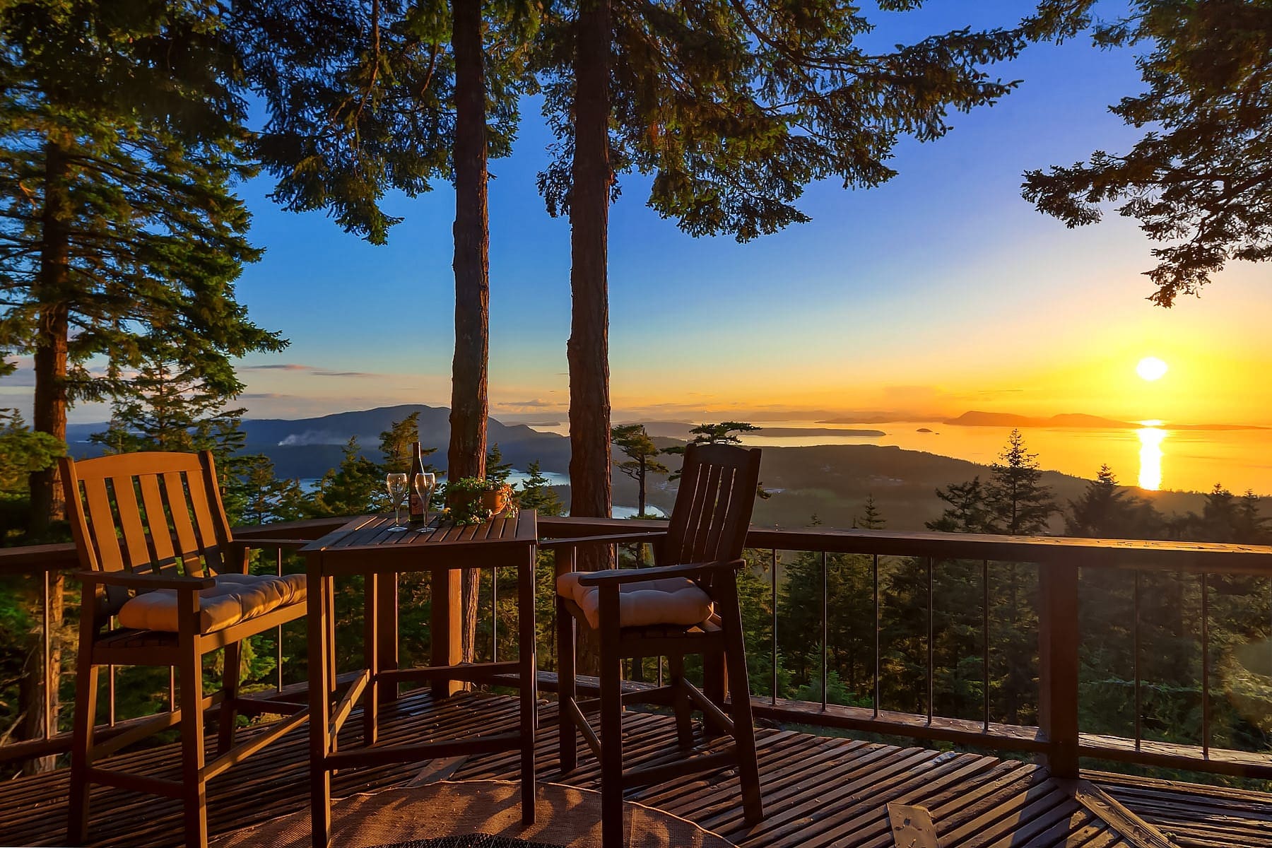 A wooden deck with a small table and two chairs offers a scenic view of a sunset over the ocean. The sun is near the horizon, casting a warm golden glow over the water and distant islands. Tall trees frame the scene, adding to the tranquil and picturesque setting.