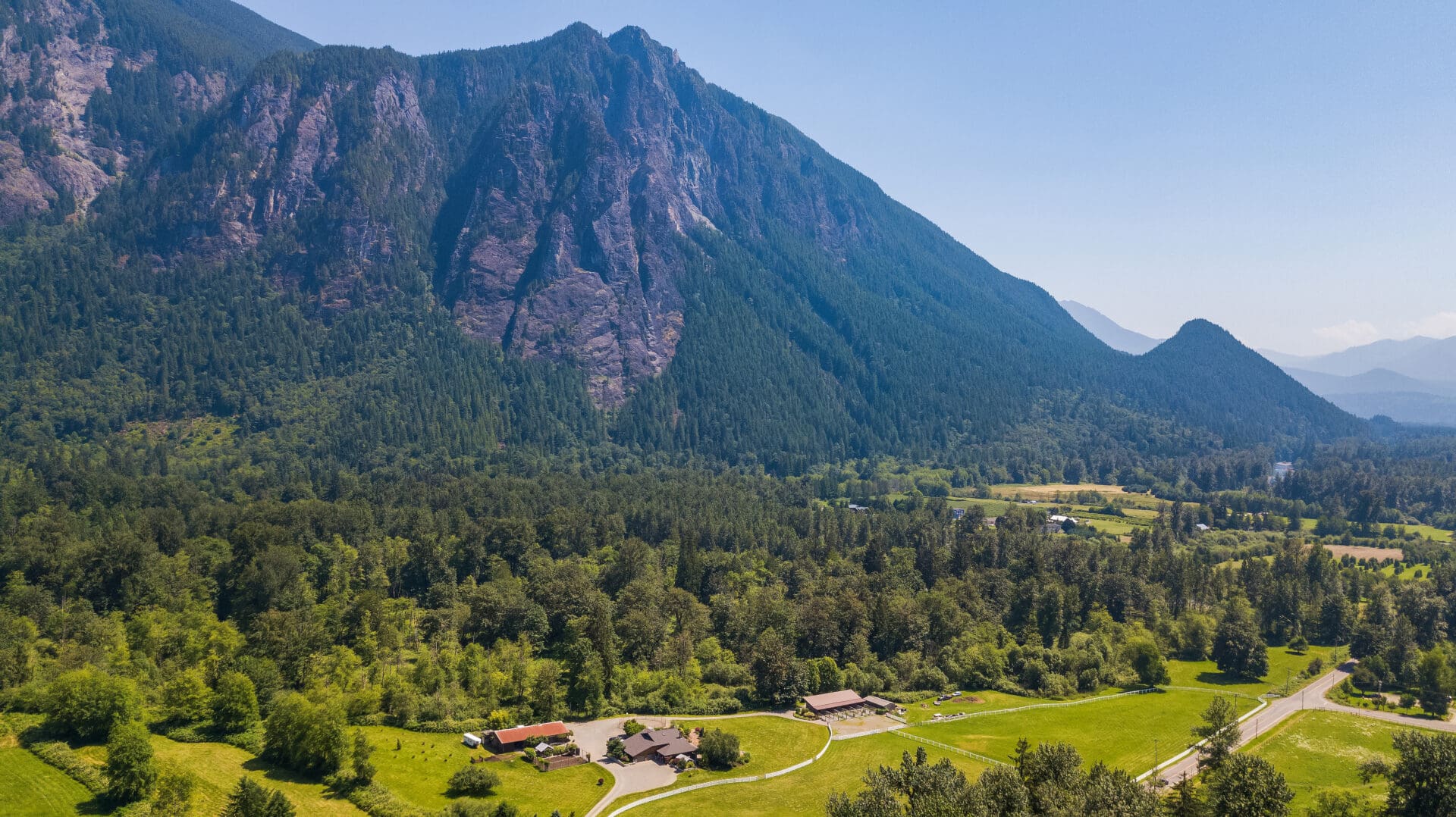 Aerial view of a picturesque landscape in the Pacific Northwest, featuring lush green fields and dense forests surrounded by majestic mountains under a clear blue sky. Ideal for showcasing rural real estate opportunities.