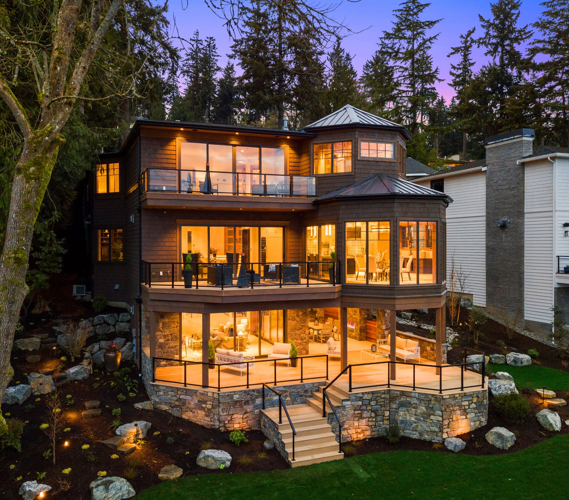 Modern exterior view of a luxury home featuring expansive windows and a stone foundation, set against a backdrop of tall trees in the Pacific Northwest. The warm evening lighting creates an inviting atmosphere.