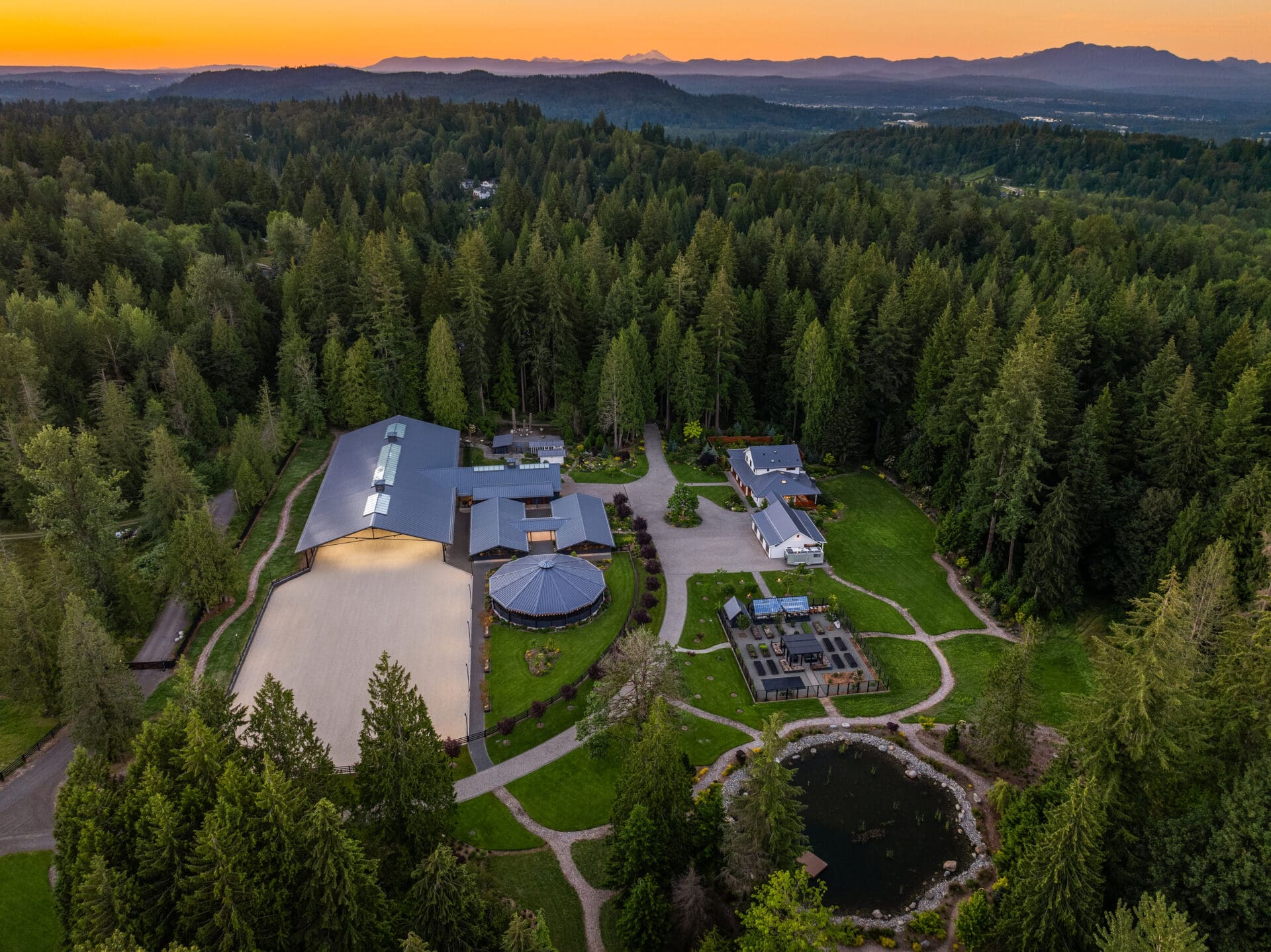 Aerial view of a luxurious estate in the Pacific Northwest, featuring modern architectural designs, expansive green lawns, and a serene pond surrounded by lush trees at twilight.
