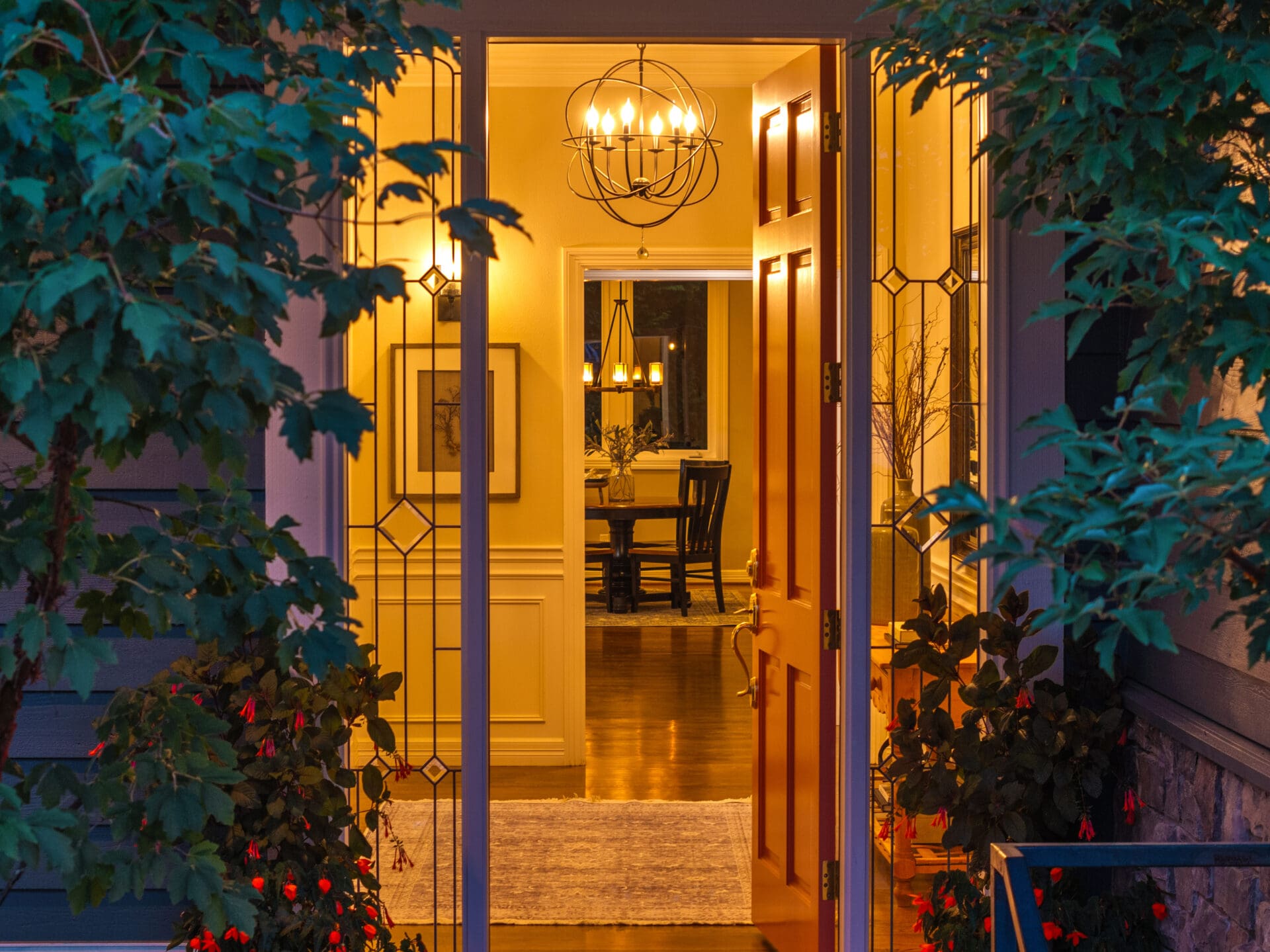 Welcoming entryway of a modern home in the Pacific Northwest, featuring elegant lighting and warm yellow tones, framed by lush greenery and vibrant flowers.
