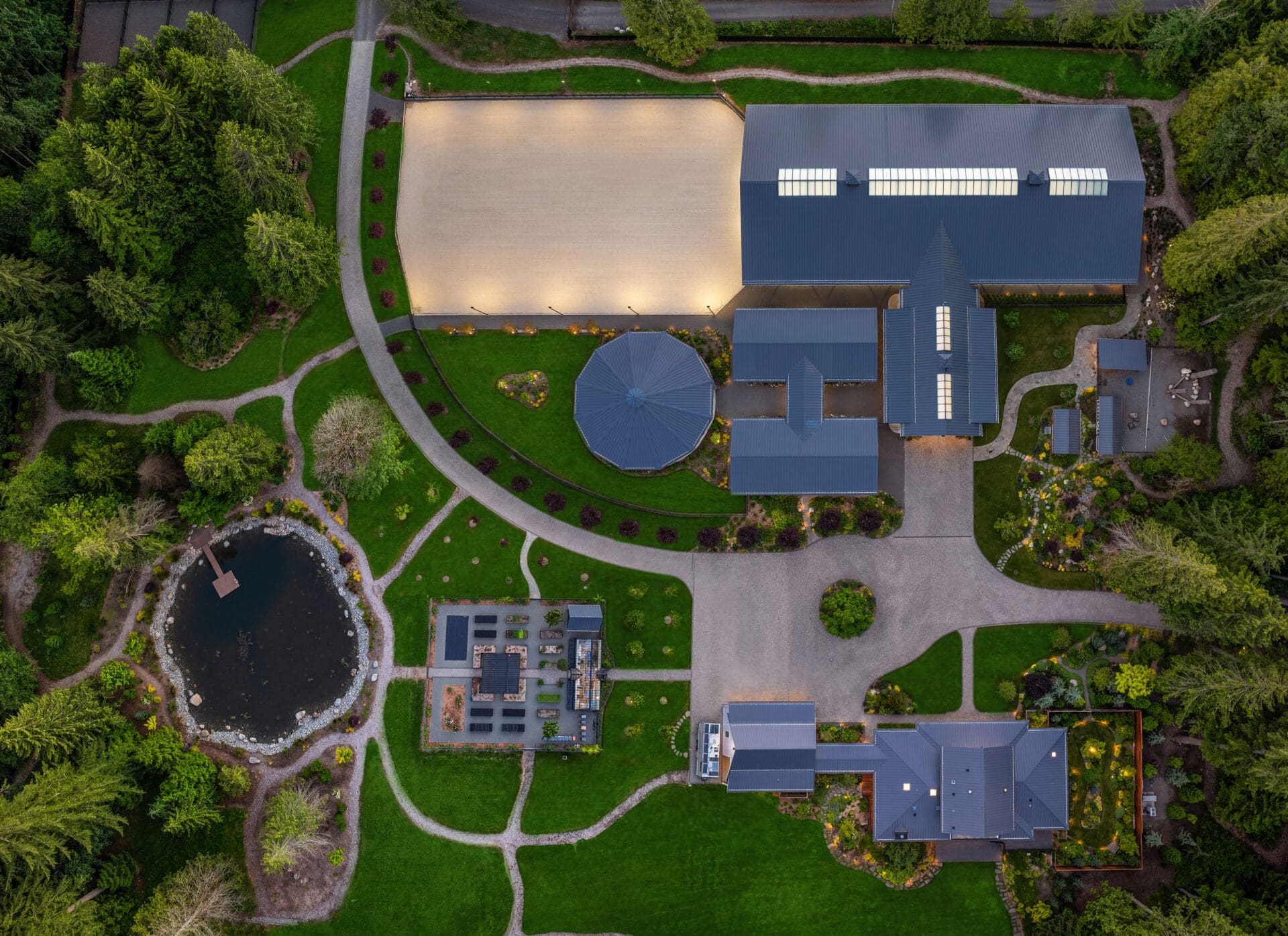Aerial view of a luxurious estate in the Pacific Northwest featuring modern architecture, manicured gardens, a pond, and a spacious riding arena, showcasing serene outdoor living spaces.