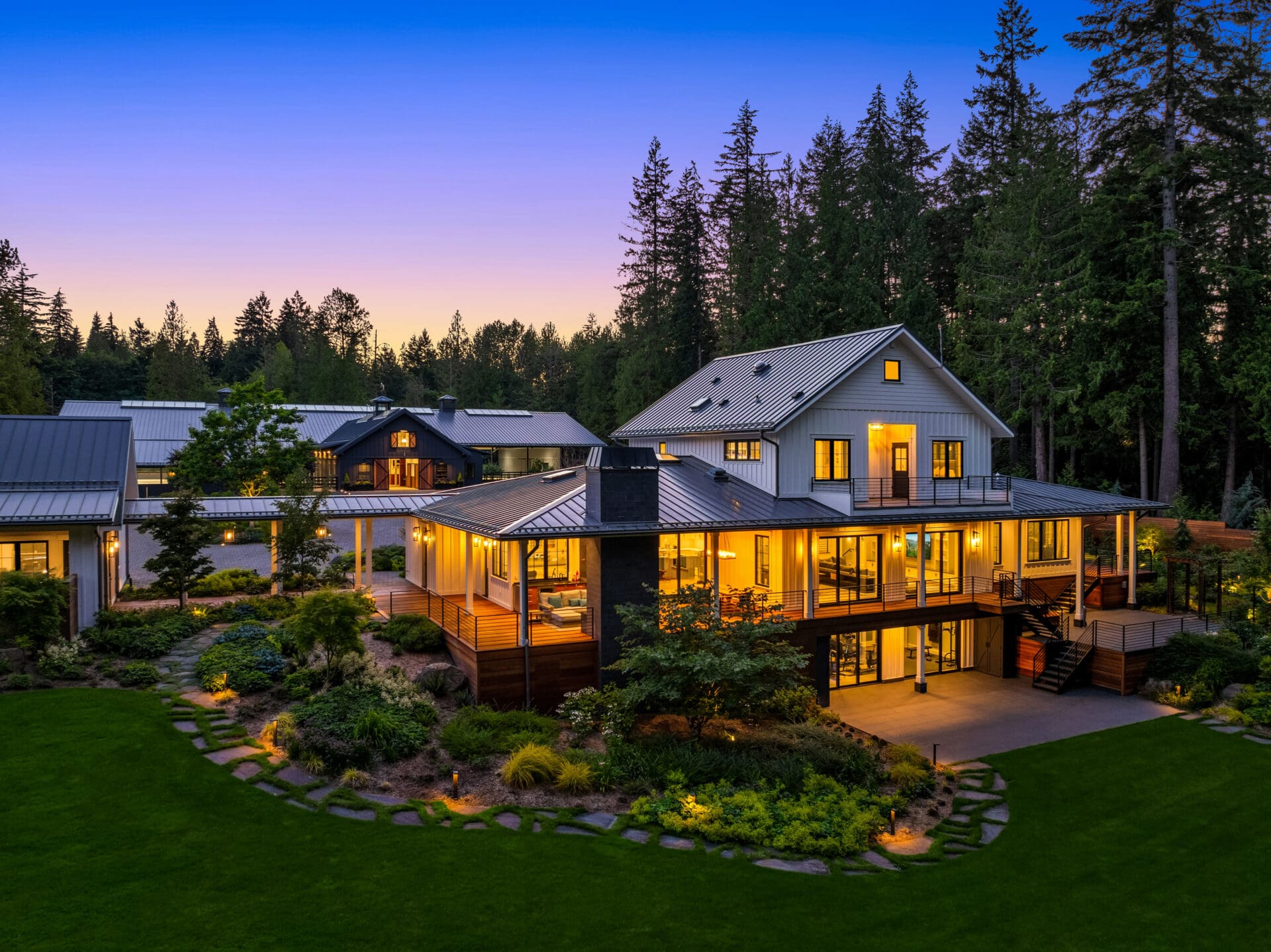 Modern exterior of a luxury home in the Pacific Northwest, featuring elegant landscaping, warm lighting, and a backdrop of towering trees at twilight.