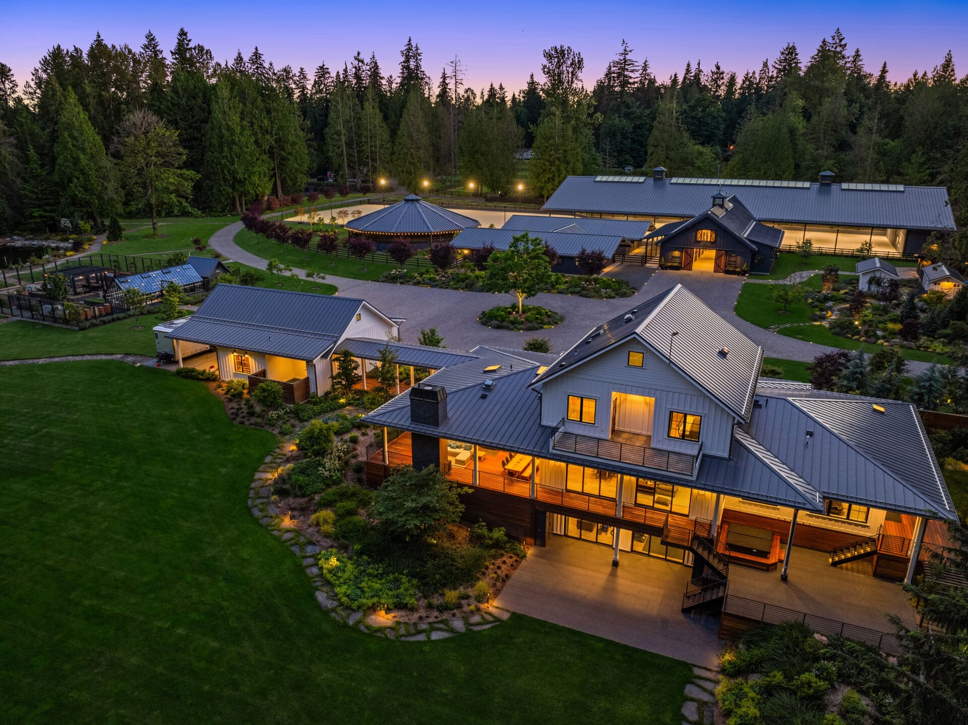 Aerial view of a modern farmhouse in the Pacific Northwest, featuring elegant landscaping, a spacious lawn, and warm outdoor lighting at twilight, showcasing a blend of natural beauty and contemporary design.