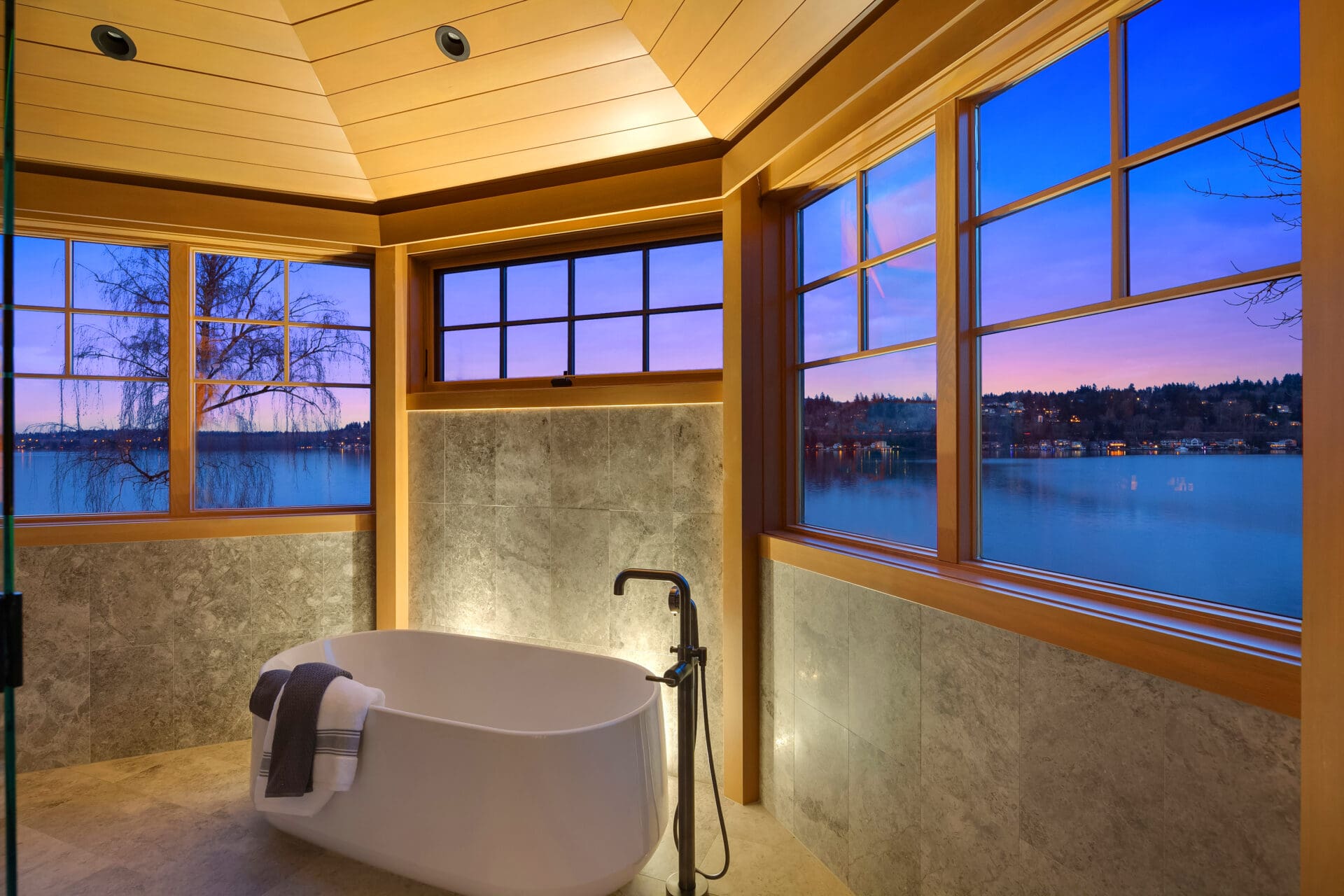 Modern bathroom with a freestanding tub and stunning lake views at twilight, featuring sleek stone walls and warm wooden accents, located in the Pacific Northwest.