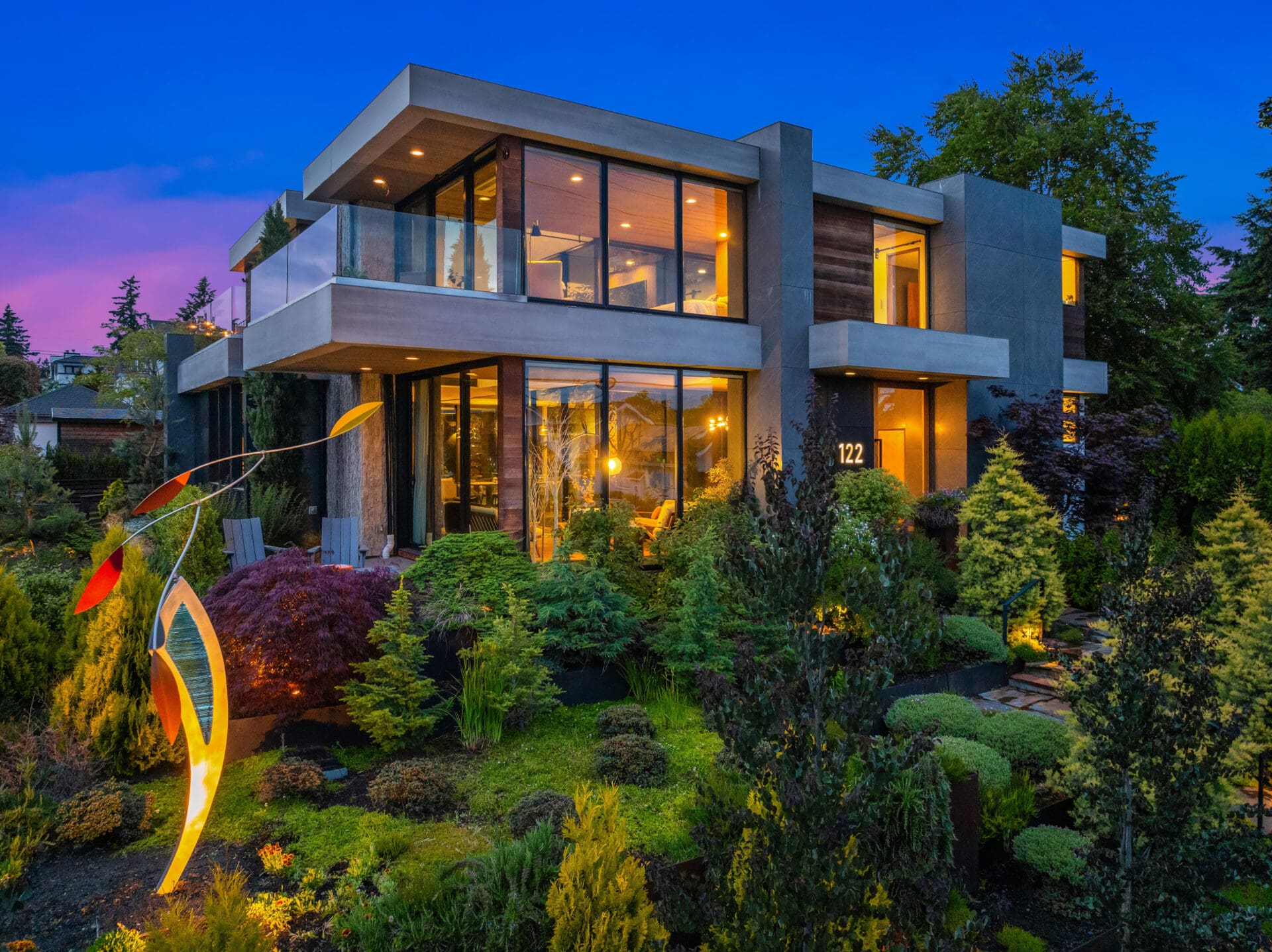 Modern exterior of a luxurious home in the Pacific Northwest, featuring large glass windows, a stylish entrance, and beautifully landscaped gardens, illuminated at twilight.