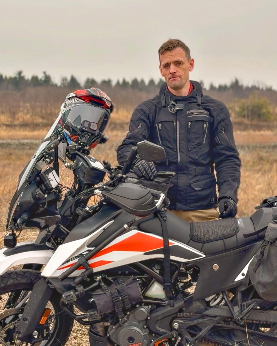 This image features a motorcyclist in a rugged outdoor setting, wearing a black riding jacket and standing next to a dual-sport motorcycle. The background showcases a muted landscape with sparse vegetation, conveying a sense of adventure and exploration. Ideal for outdoor enthusiasts in the Pacific Northwest.