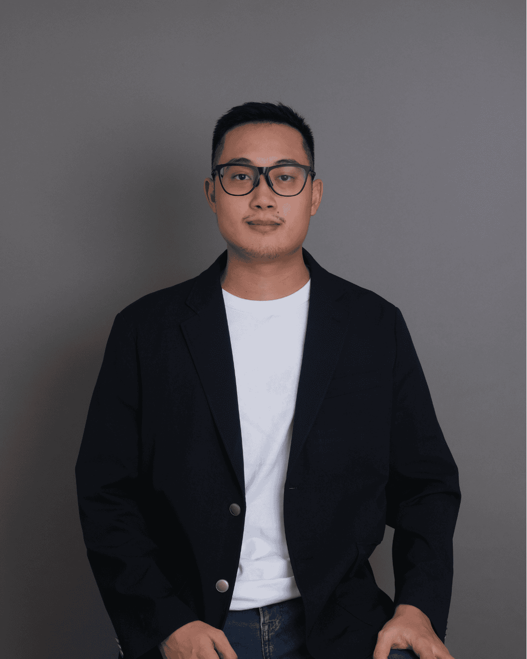 A professional portrait of a young man in a black blazer and white t-shirt against a neutral gray background, showcasing a modern and sophisticated style, ideal for real estate branding in Seattle.