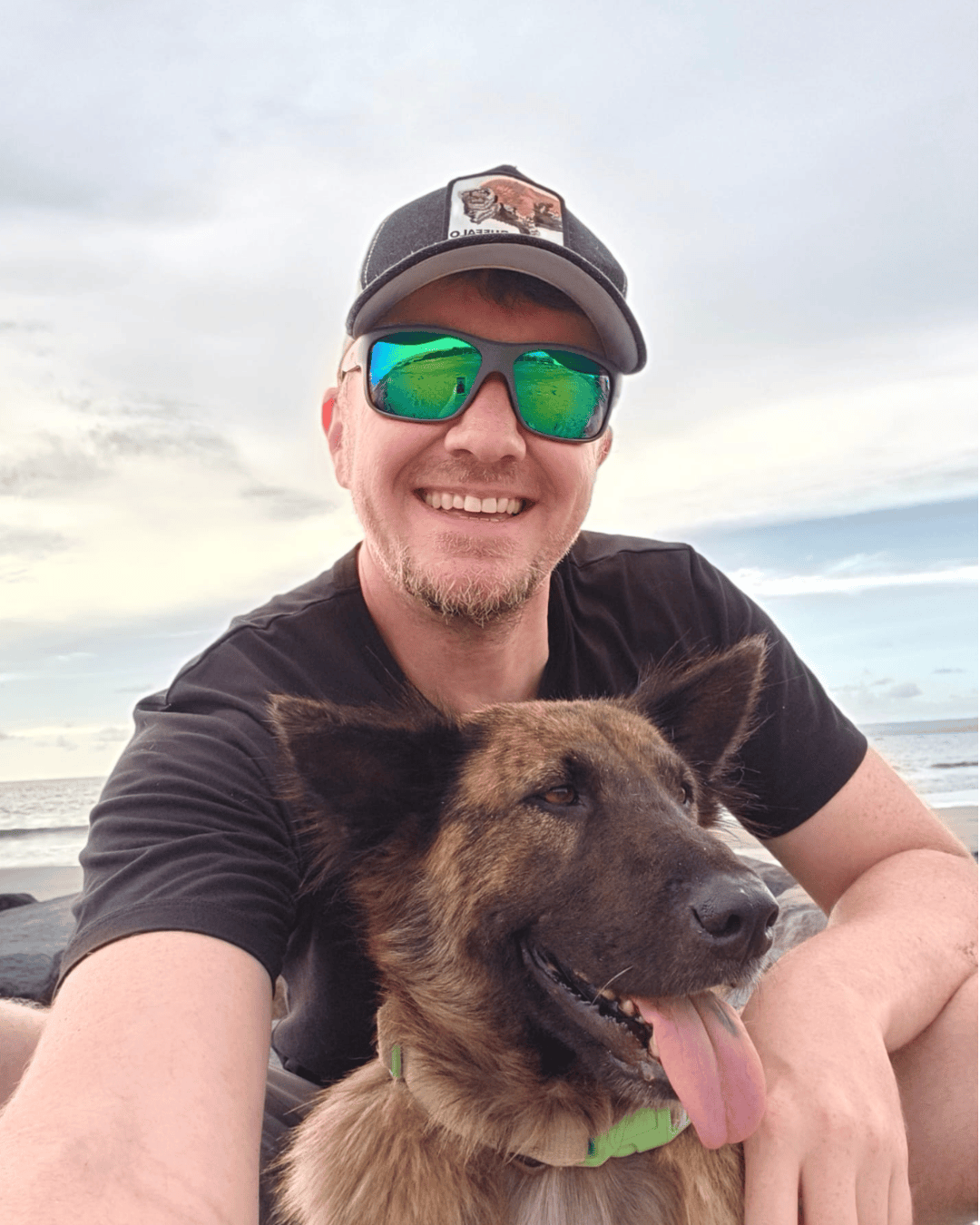 A man with sunglasses and a cap smiling with his dog on a beach at sunset in the Pacific Northwest.