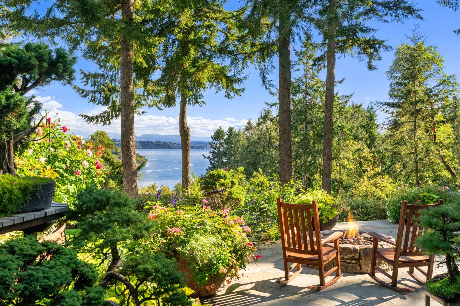 Scenic outdoor patio with two wooden rocking chairs around a fire pit, surrounded by vibrant flowers and lush greenery, offering a stunning view of the water and trees in Seattle, Washington.