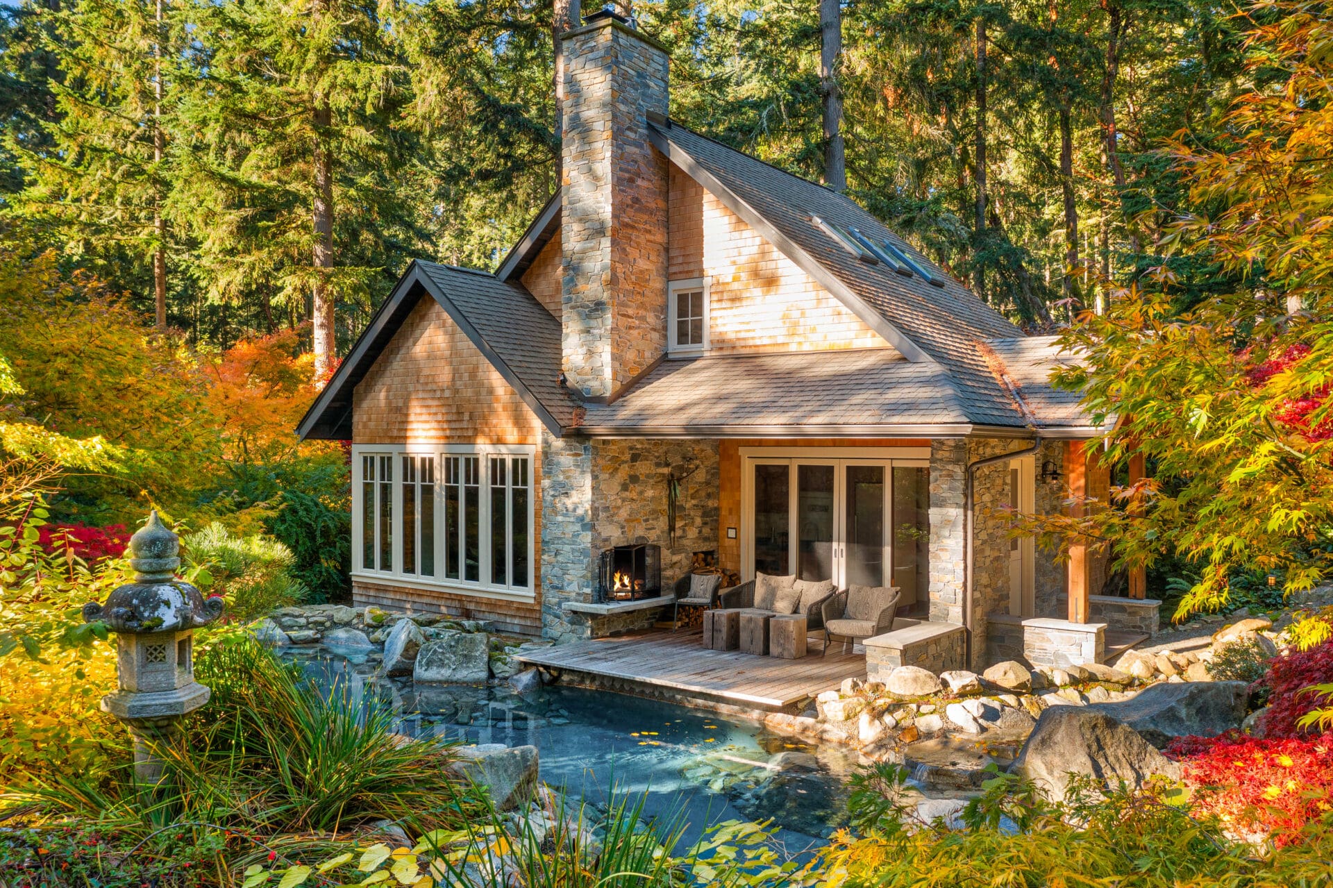 Charming exterior of a modern cabin surrounded by vibrant autumn foliage in the Pacific Northwest, featuring a stone chimney, large windows, and a cozy outdoor seating area by a tranquil pond.