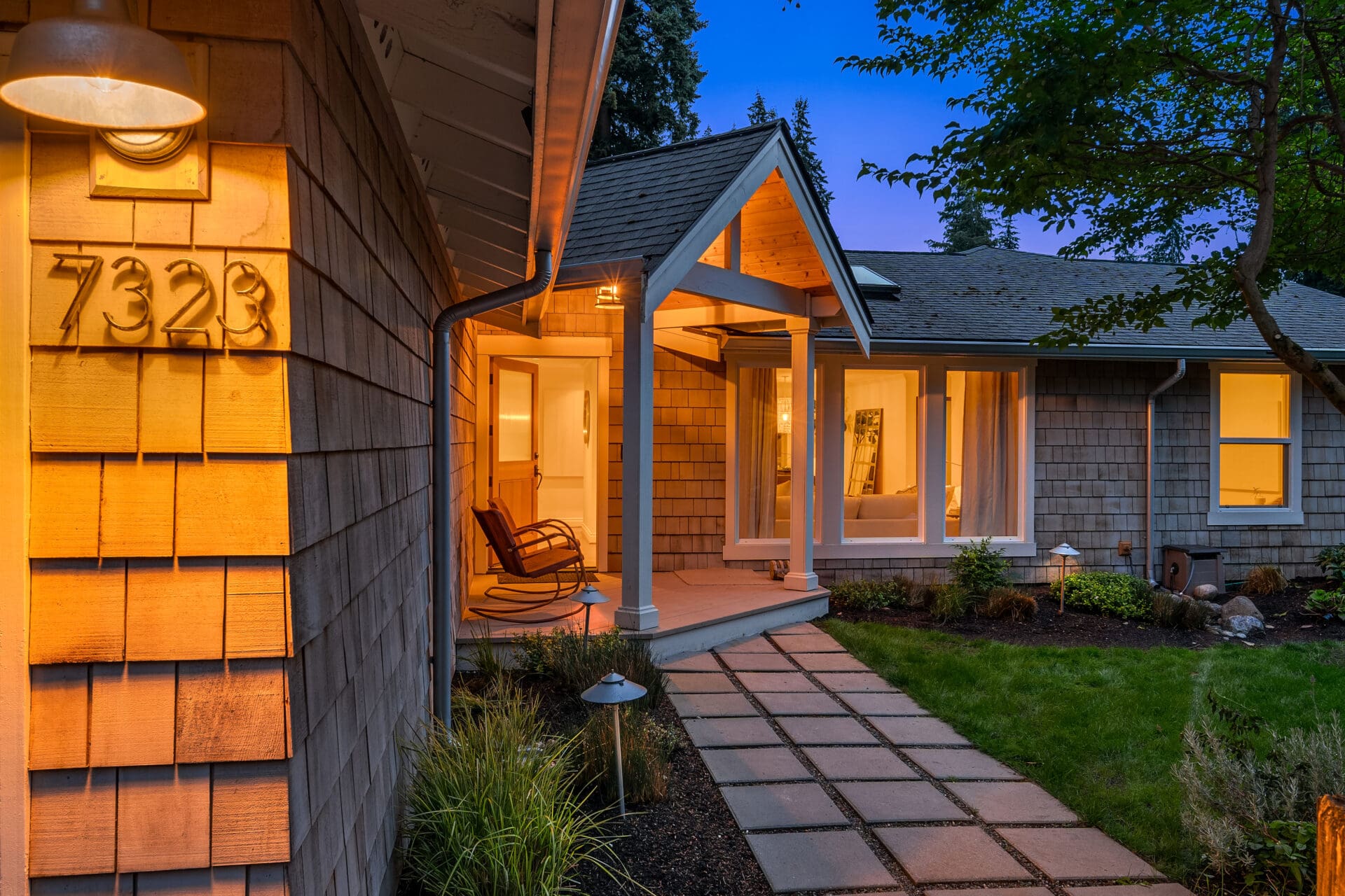 Charming exterior entrance of a home in the Pacific Northwest, featuring warm lighting, a welcoming porch with a rocking chair, and well-maintained landscaping, creating an inviting twilight ambiance.