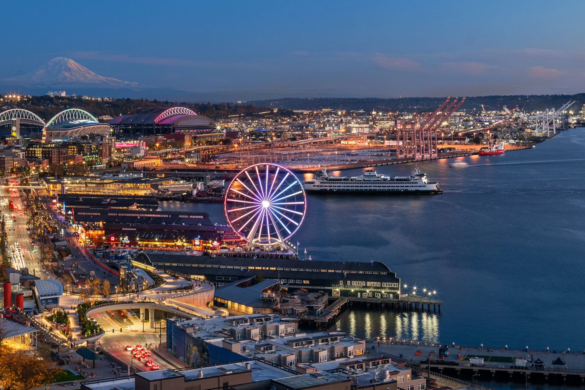 Panoramic view of Seattle's waterfront at twilight, featuring the iconic Seattle Great Wheel, vibrant city lights, and Mount Rainier in the background, showcasing the lively atmosphere of the Pacific Northwest.
