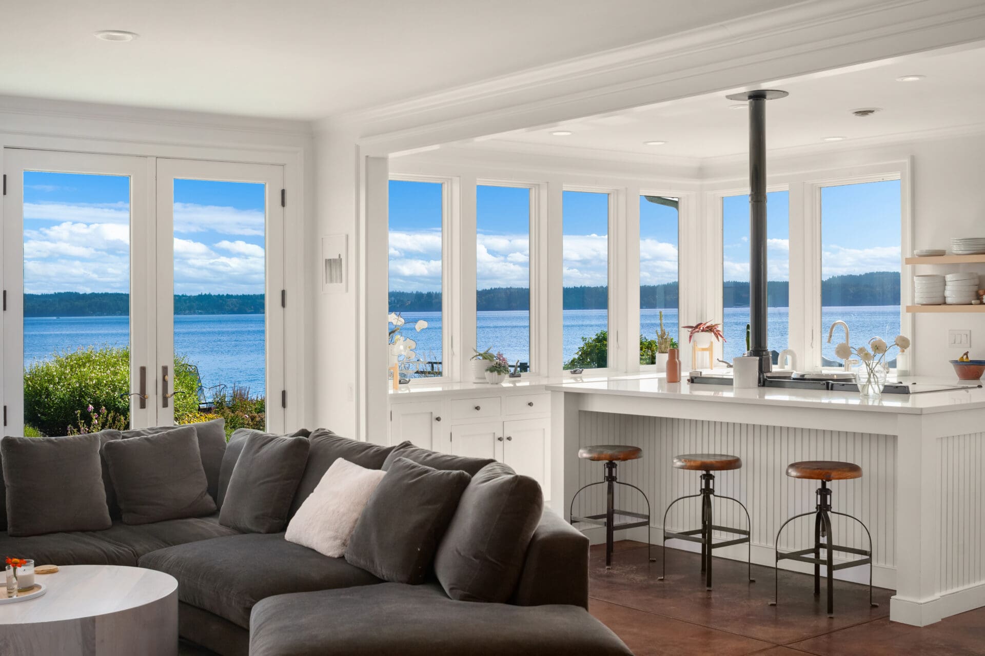 Modern kitchen with panoramic waterfront views, featuring sleek white cabinetry, a central island with bar stools, and large windows that flood the space with natural light. Located in the Pacific Northwest.