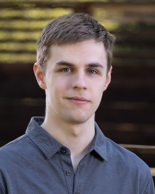 Young man with short hair wearing a gray polo shirt, standing outdoors with a blurred wooden background in a natural setting, Seattle.