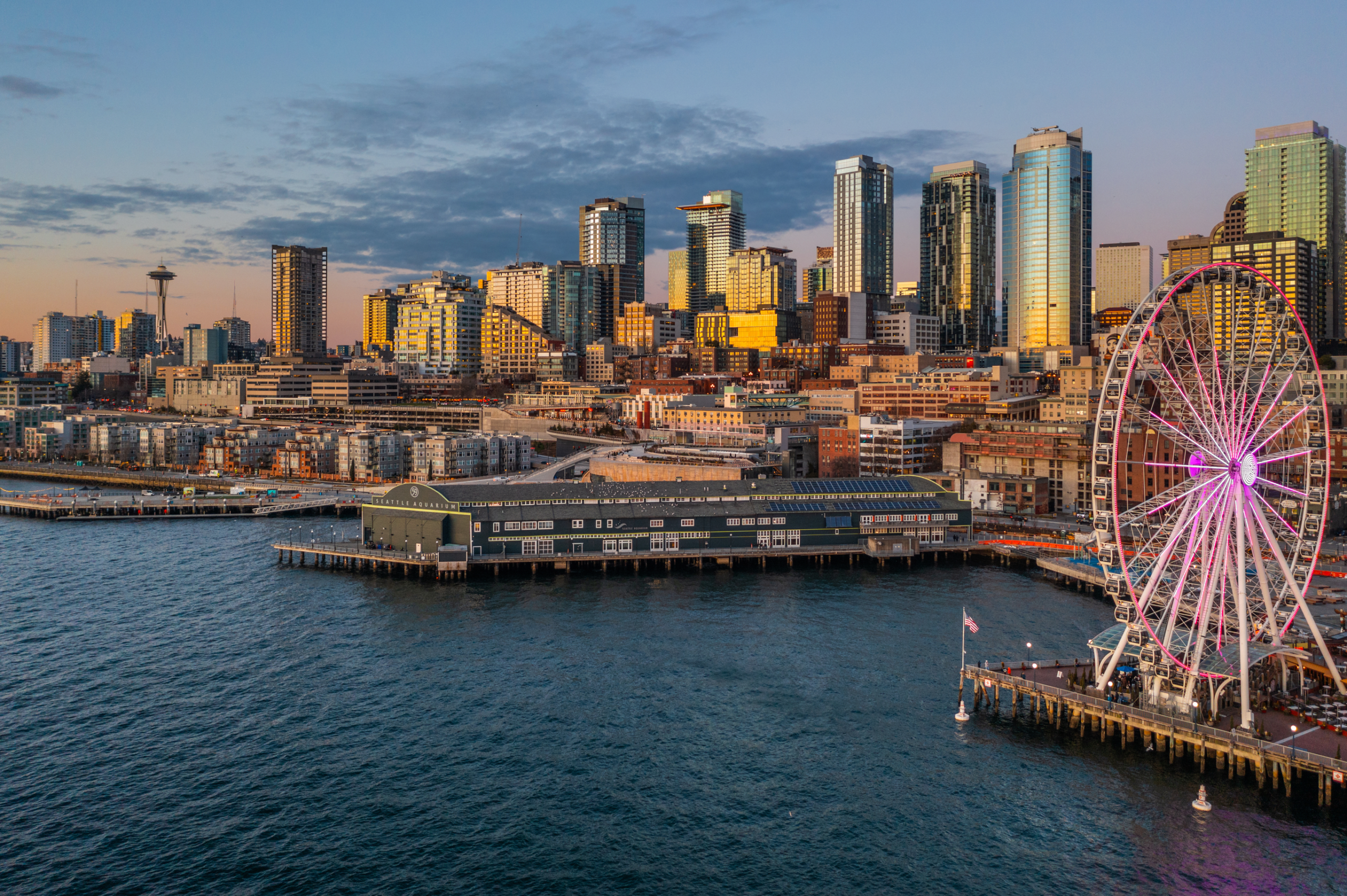 Seattle waterfront skyline at twilight featuring modern high-rise buildings, the iconic Space Needle, and the Seattle Great Wheel illuminated, showcasing a vibrant urban atmosphere in the Pacific Northwest.