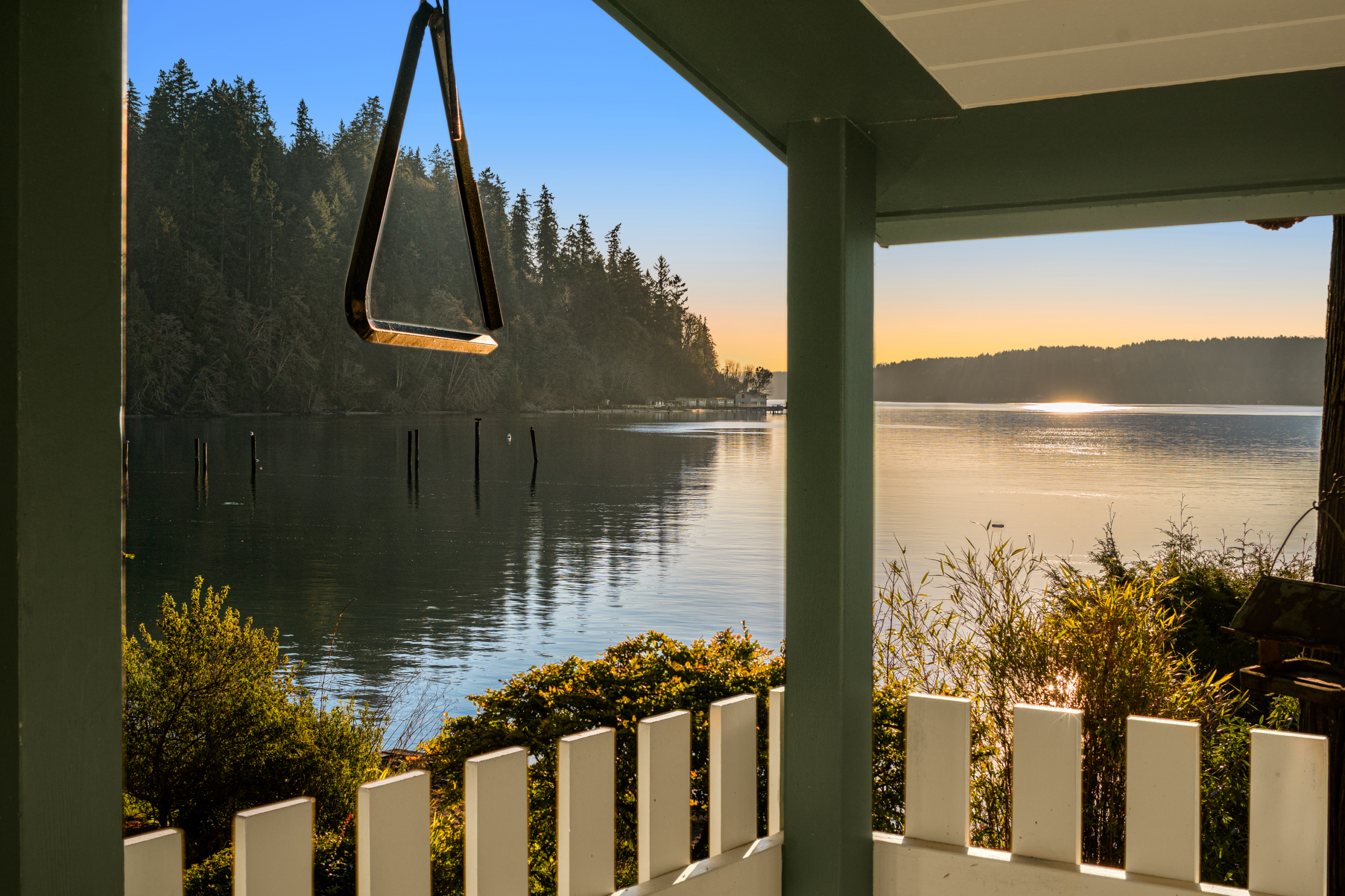 Scenic waterfront view from a cozy porch, featuring a swing and lush greenery, with a tranquil lake reflecting the sunset in the Pacific Northwest.
