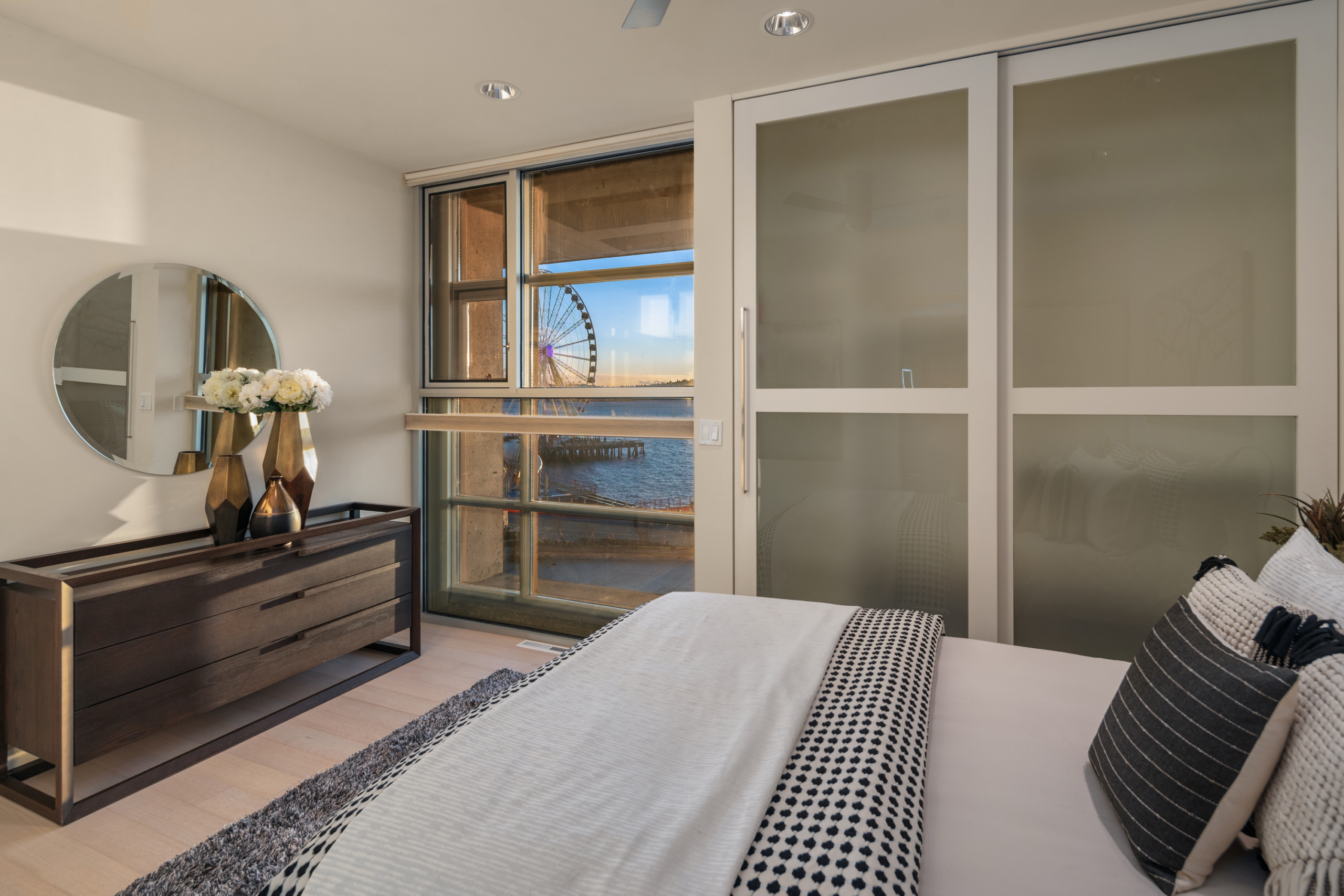 Modern bedroom with sleek furnishings and large windows showcasing a waterfront view and Seattle's iconic Ferris wheel. The space features neutral tones, stylish decor, and abundant natural light, creating a serene atmosphere.