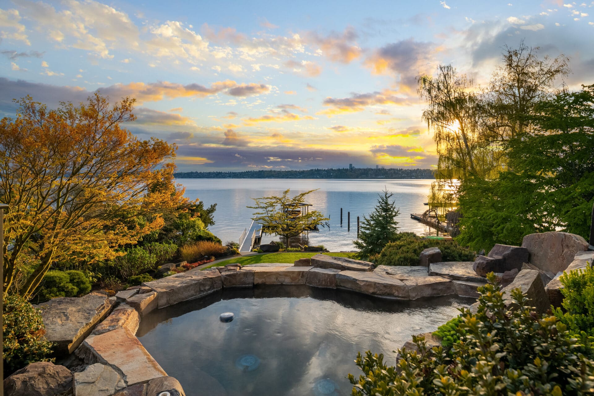 Stunning waterfront view from a beautifully landscaped backyard featuring a stone hot tub, lush greenery, and vibrant autumn trees, set against a picturesque Seattle sunset.