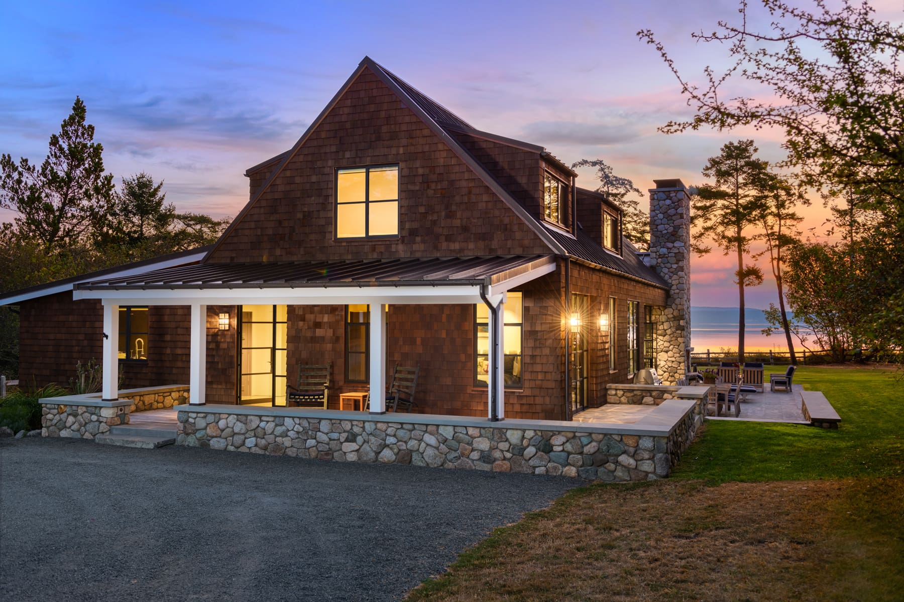 Charming exterior of a modern rustic home at twilight, featuring wood shingles and stone accents, set against a serene waterfront backdrop in the Pacific Northwest.