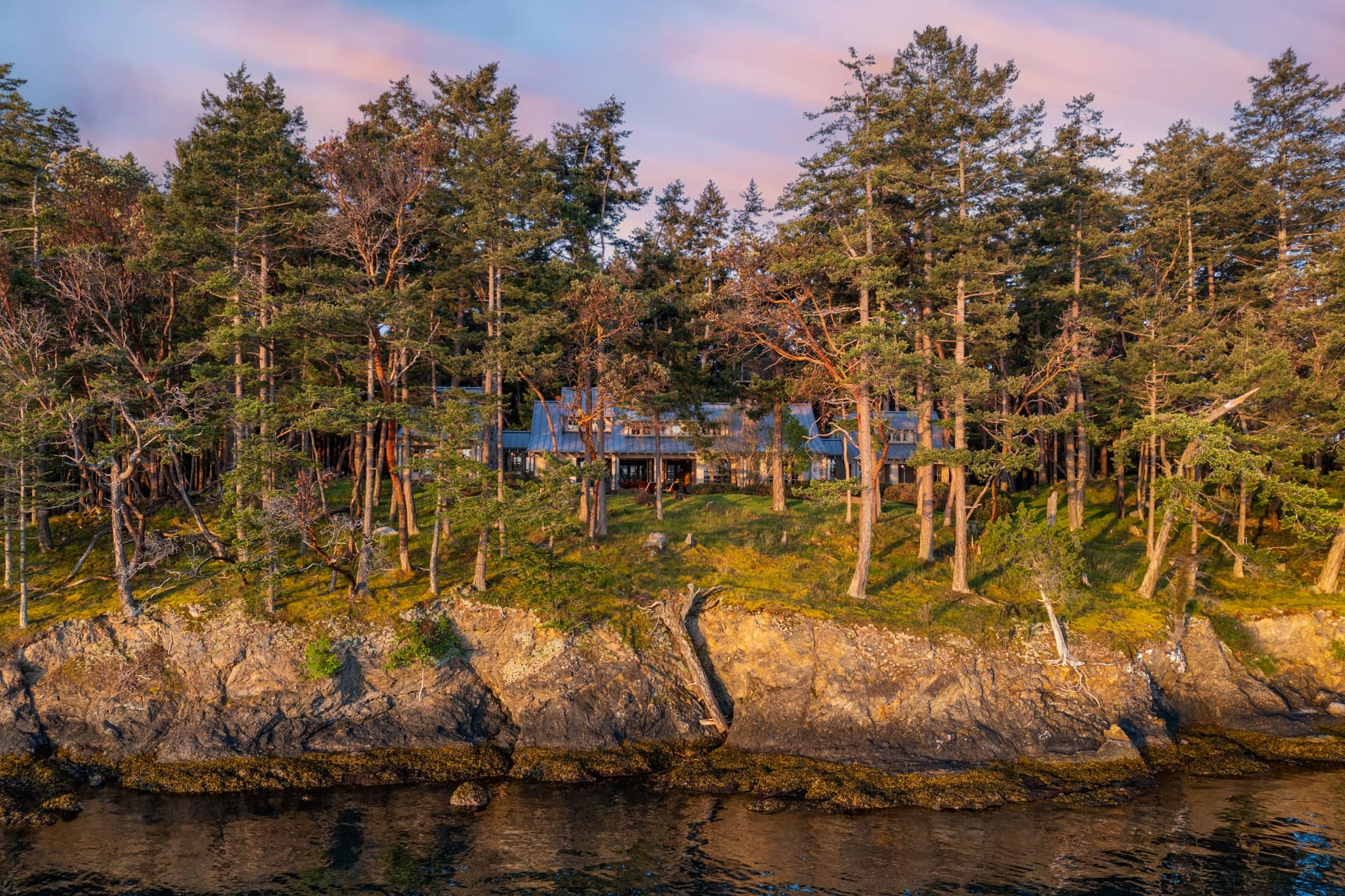 Stunning waterfront property nestled among trees on a rocky shoreline in the Pacific Northwest, showcasing a modern home with large windows reflecting the serene natural surroundings.