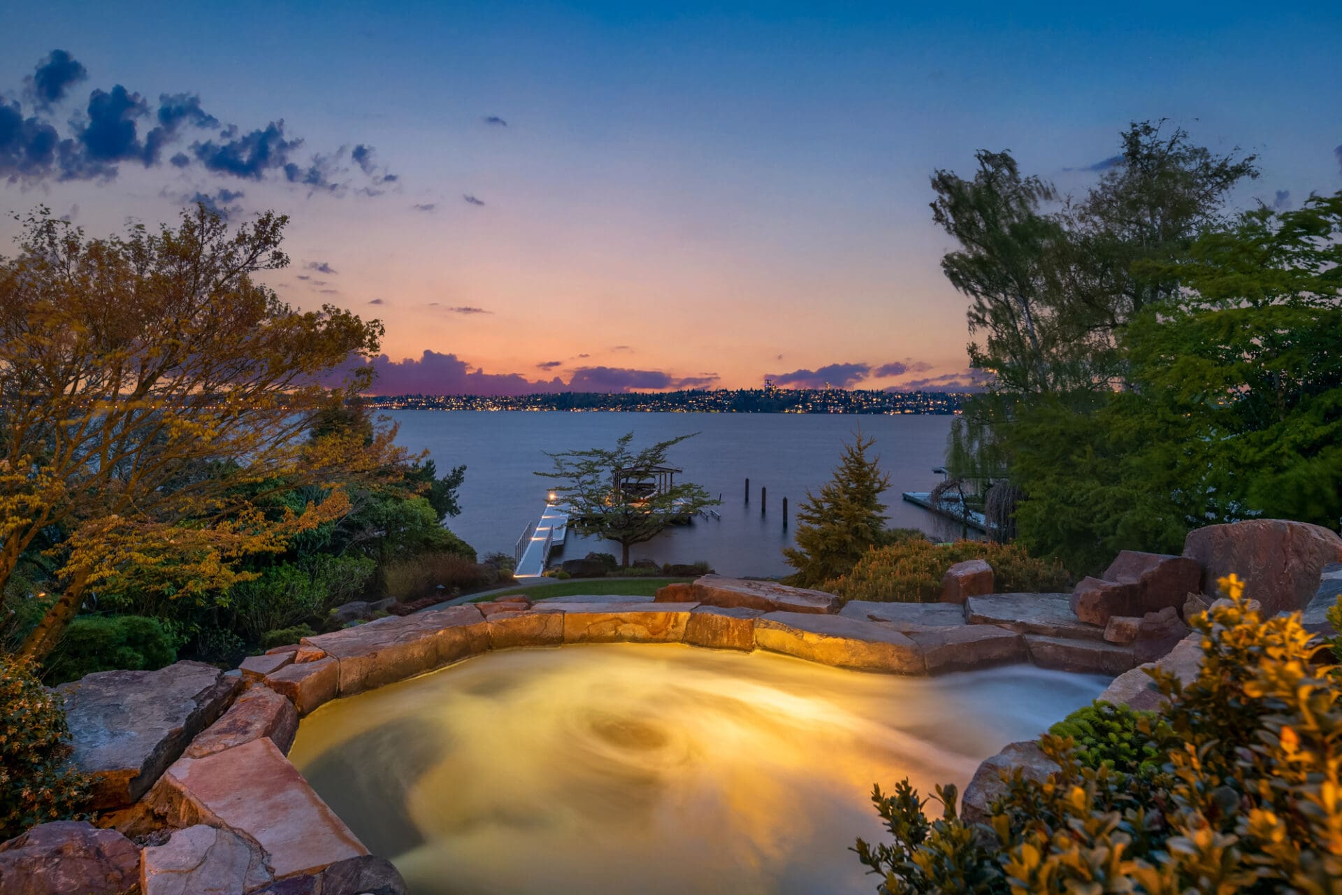 Scenic waterfront hot tub with natural stone surround, overlooking a tranquil lake at twilight, showcasing vibrant sunset colors in the Pacific Northwest.