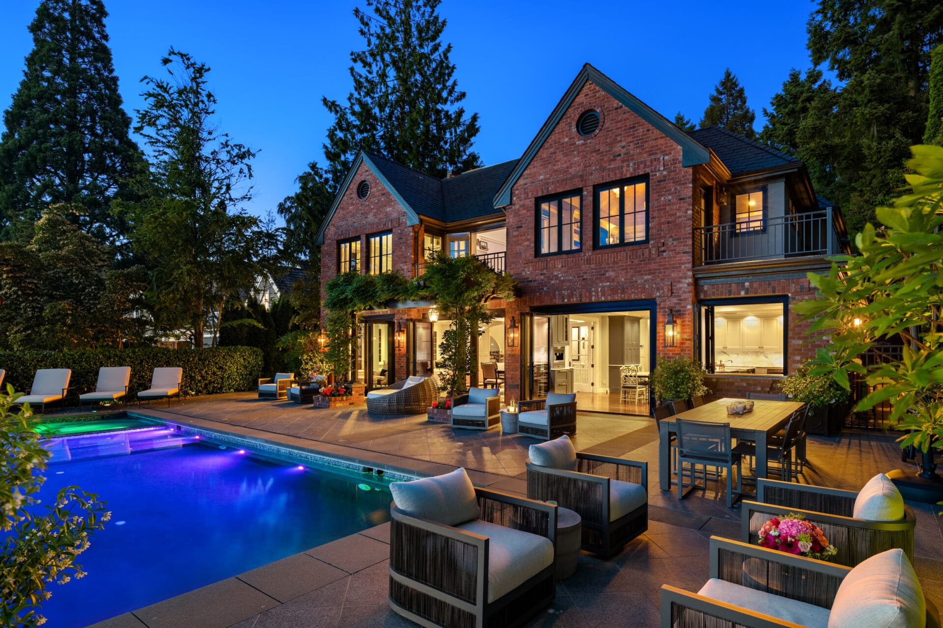 Elegant exterior of a luxury home in the Pacific Northwest, featuring a modern patio with a pool, stylish seating, and lush landscaping, illuminated at twilight.