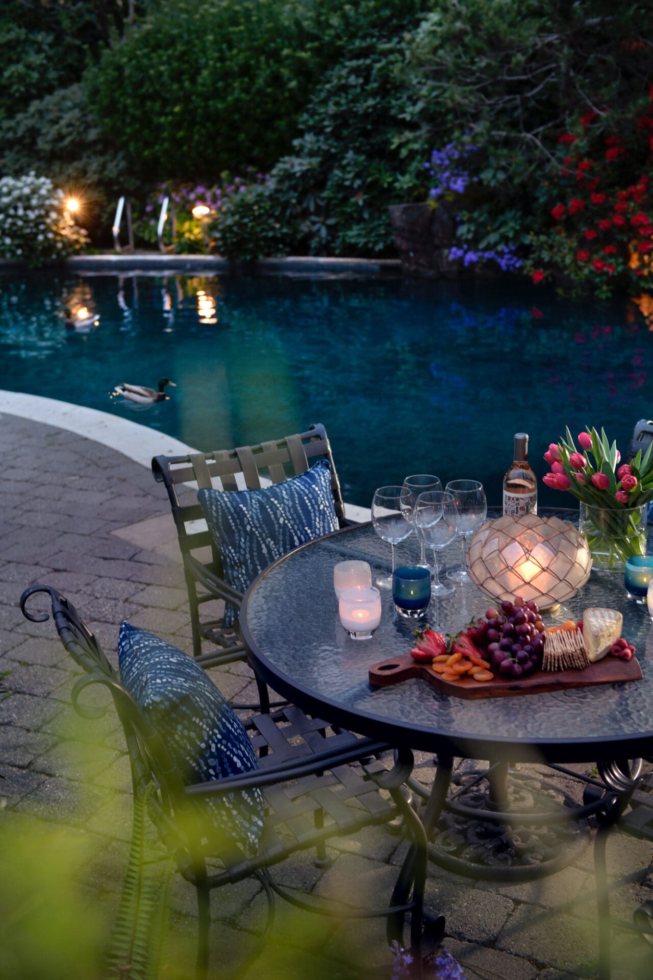 Outdoor dining area with a glass table set for a cozy evening, featuring elegant lighting, a charcuterie board, and floral accents, surrounded by a lush garden and pool in the Pacific Northwest.