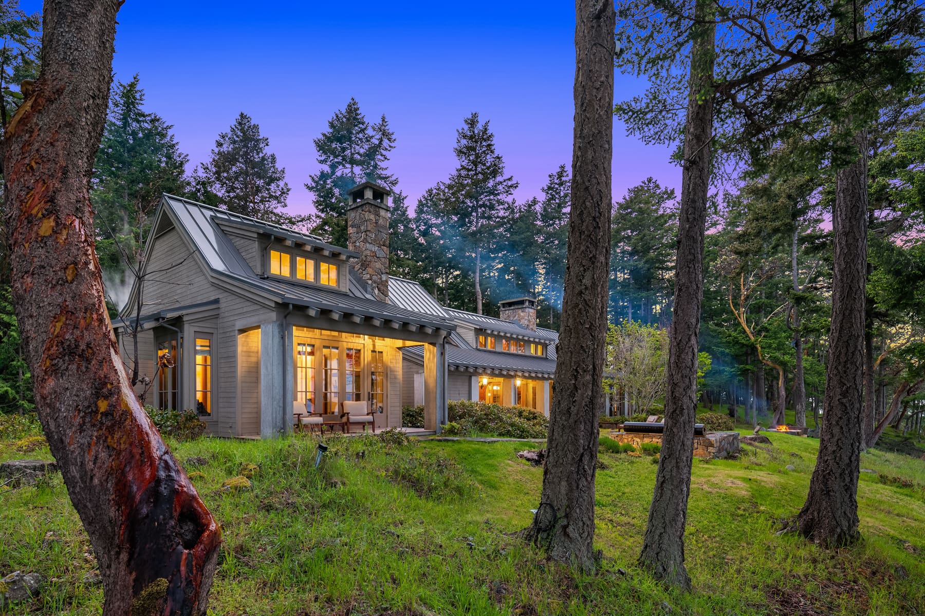 Charming exterior of a modern cottage surrounded by lush greenery in the Pacific Northwest, featuring a stone chimney and warm glowing windows against a twilight sky.