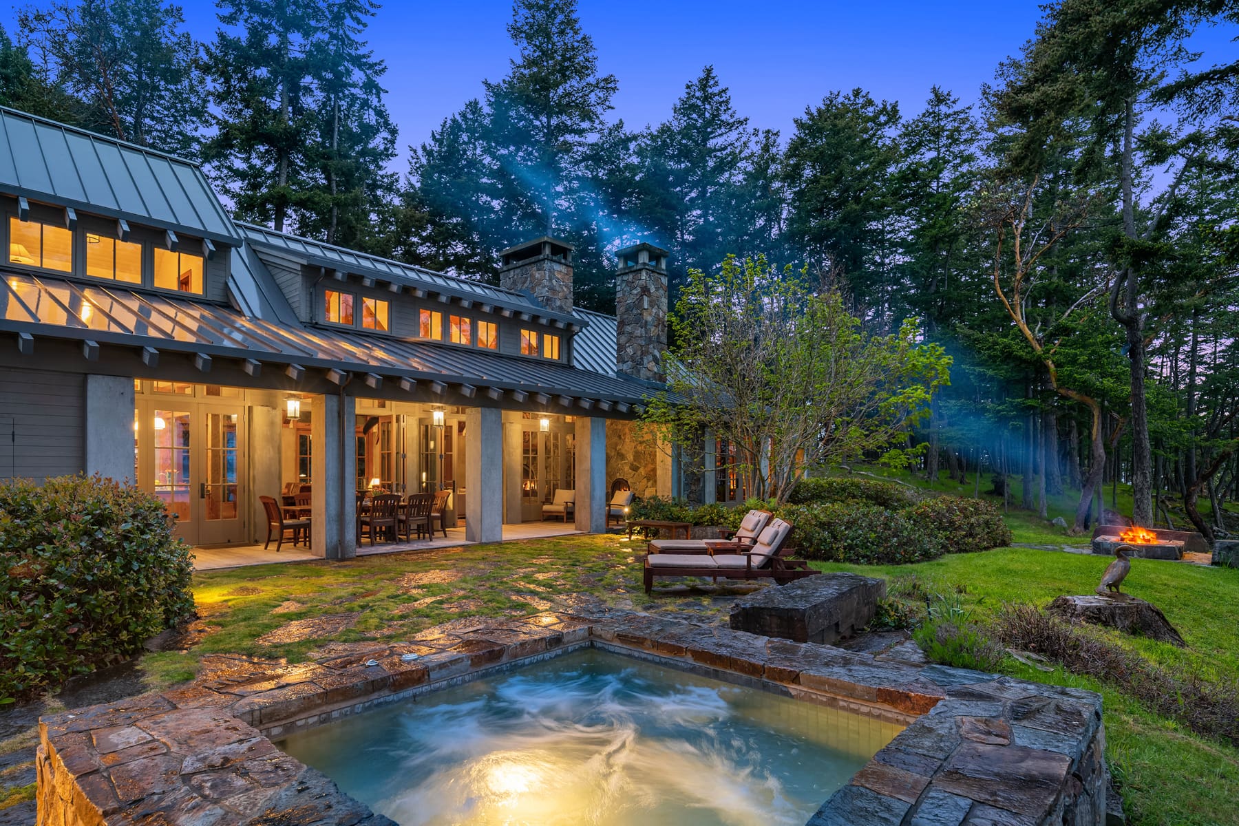 Elegant exterior of a modern home featuring a stone patio, hot tub, and outdoor dining area, surrounded by lush greenery in the Pacific Northwest at twilight.