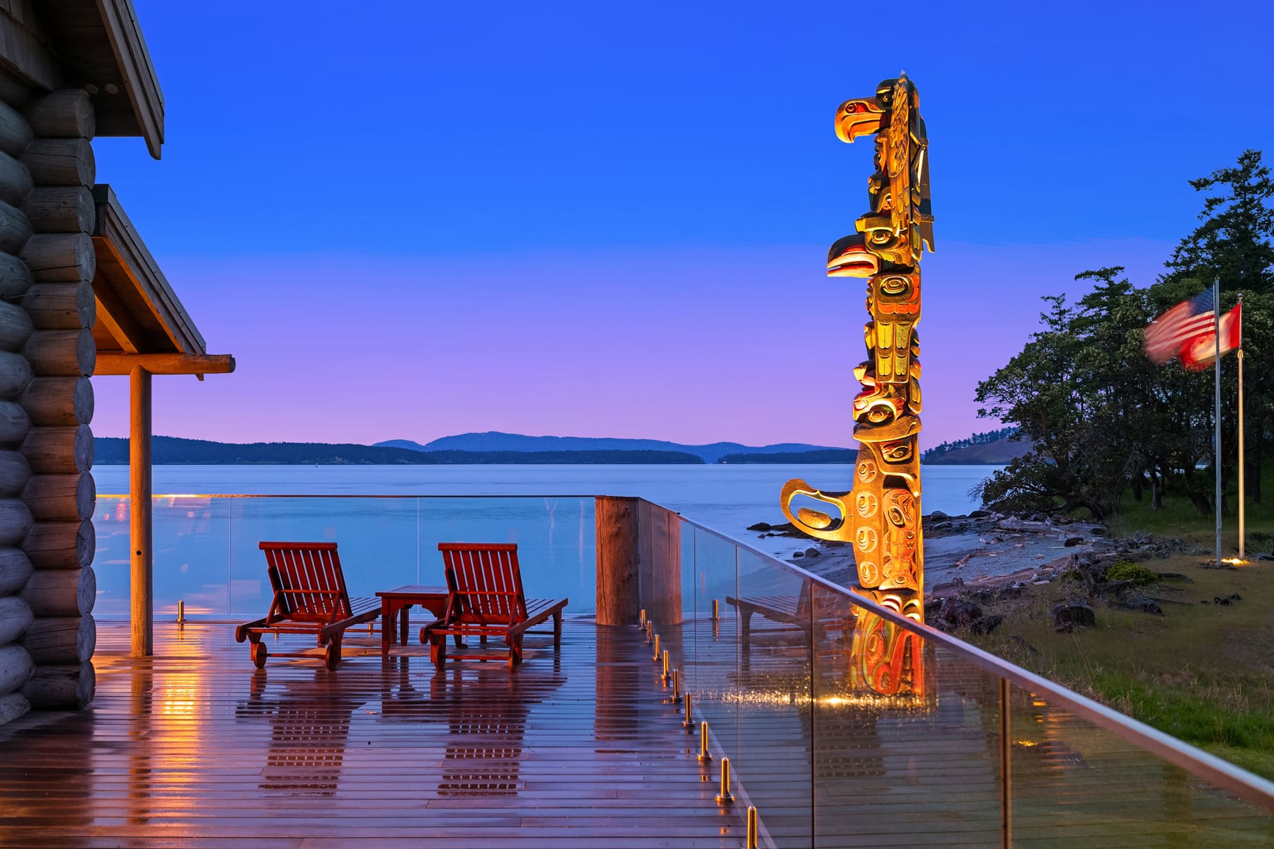 Scenic waterfront patio with red lounge chairs and a totem pole, showcasing a modern design against a twilight sky in the Pacific Northwest.